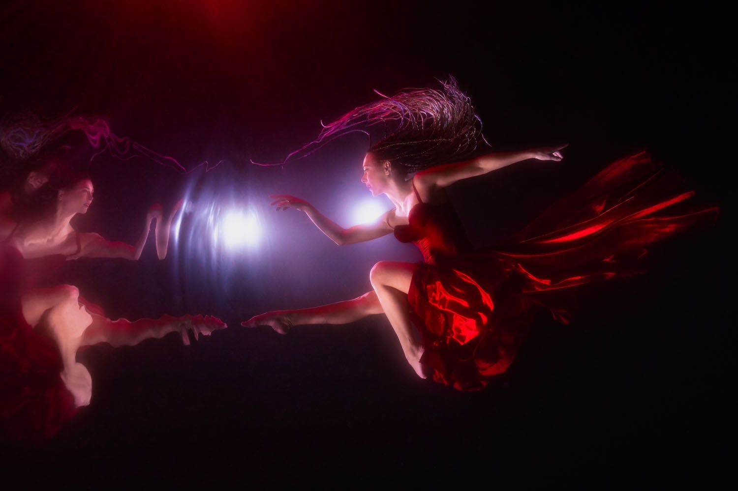 Two ethereal figures in flowing red dresses float underwater with dramatic lighting effects creating a mystical atmosphere.