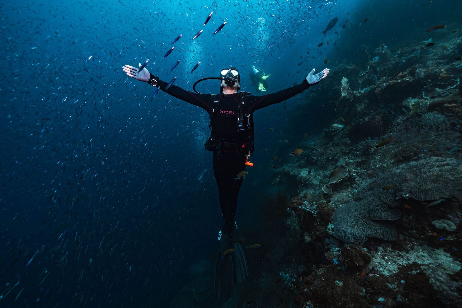 Scuba diver with arms spread wide beneath sunlit blue waters near a coral reef wall.