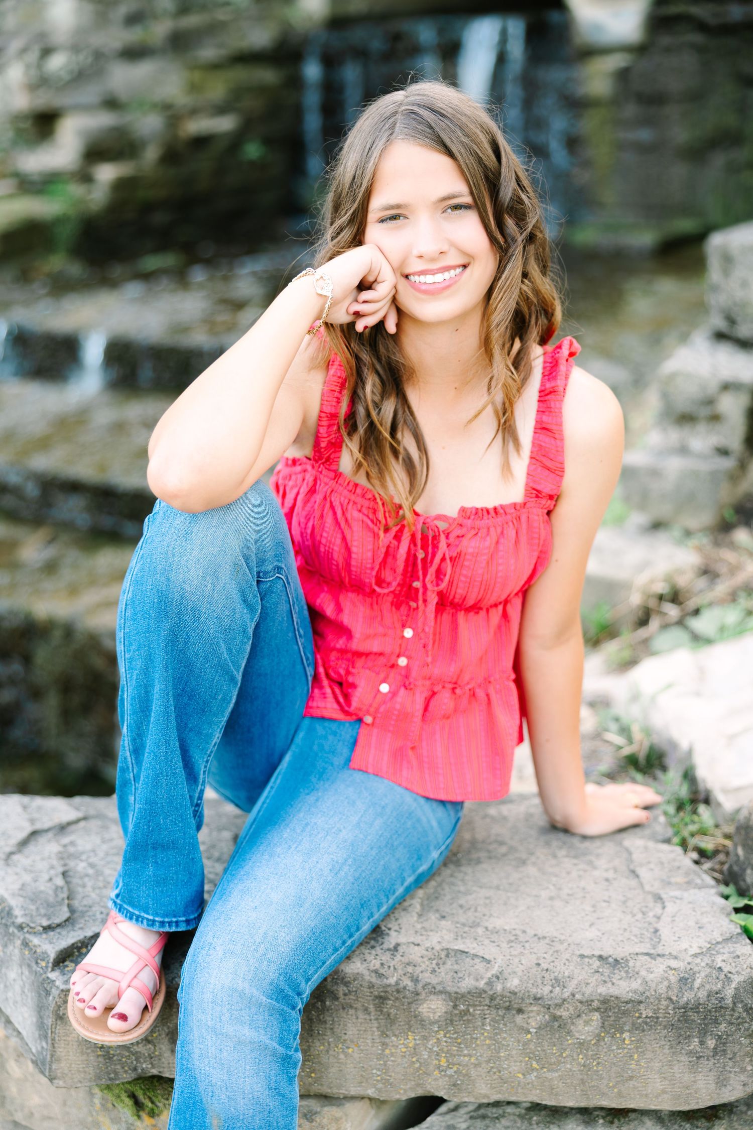 Person in red top and blue jeans sitting on rocks outdoors with natural scenery in background.