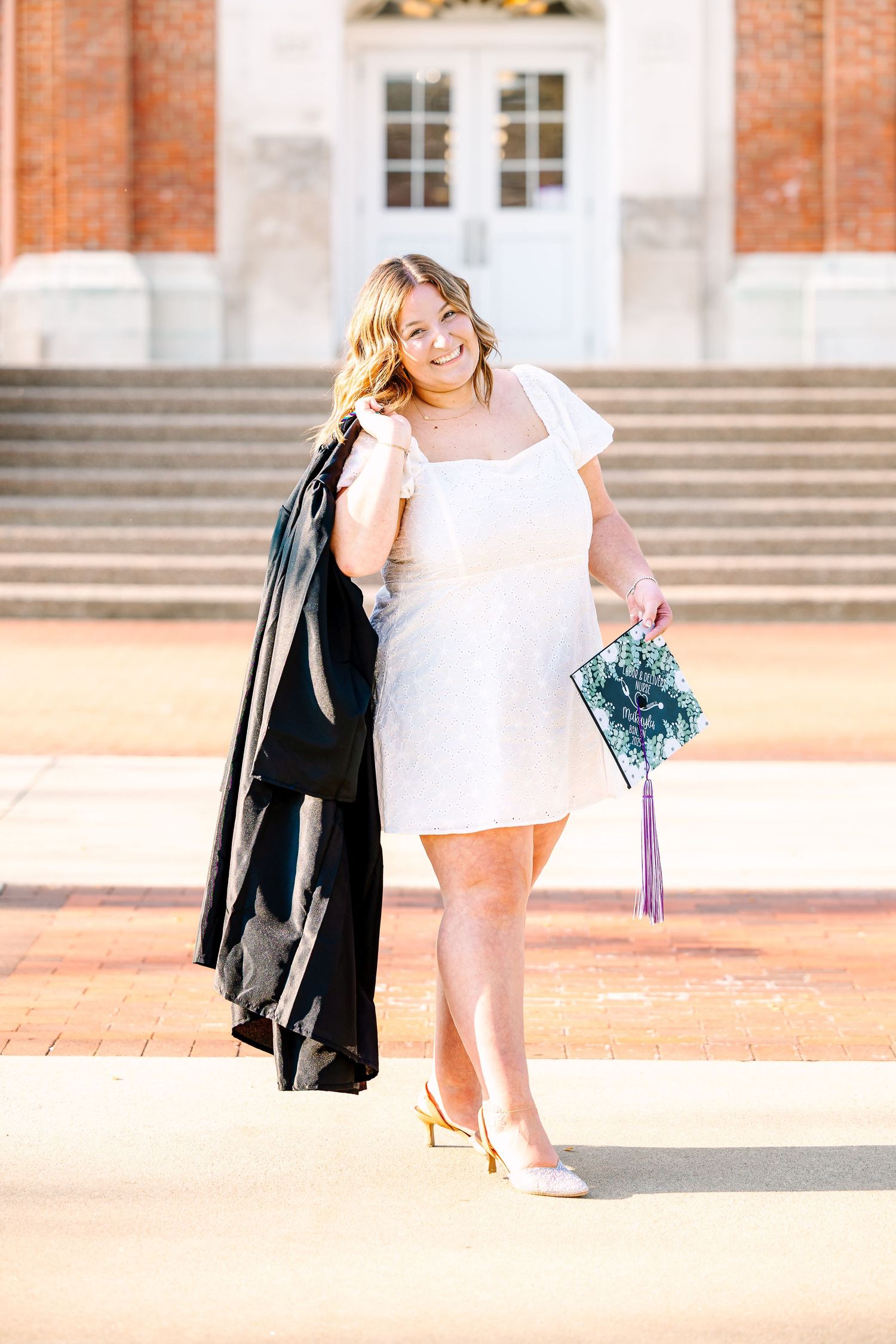 Graduation portrait of someone in a white dress carrying a black gown and blue floral bouquet on sunny campus steps.