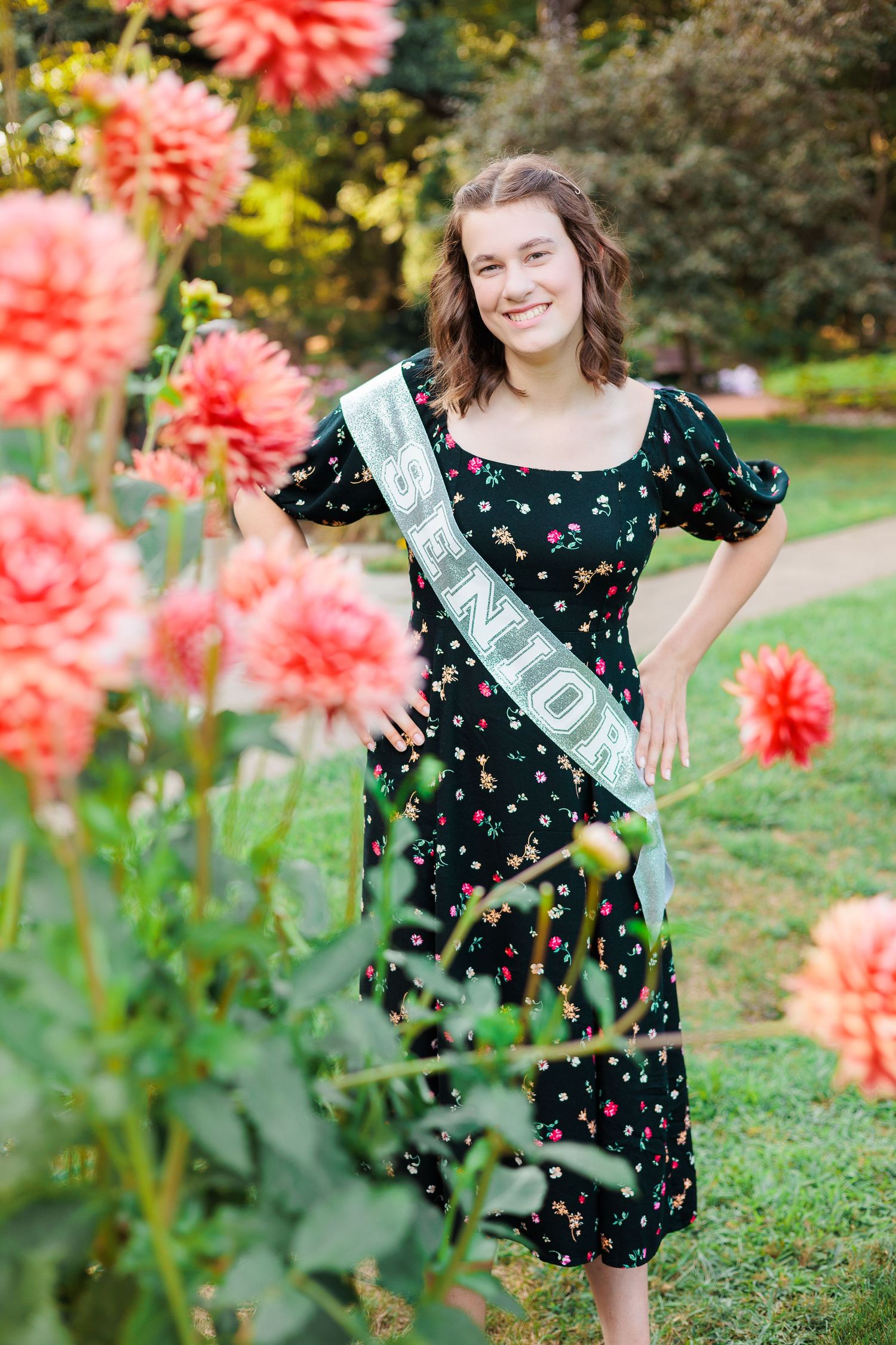 Young woman in a polka dot dress poses near bright coral dahlias in a garden setting.