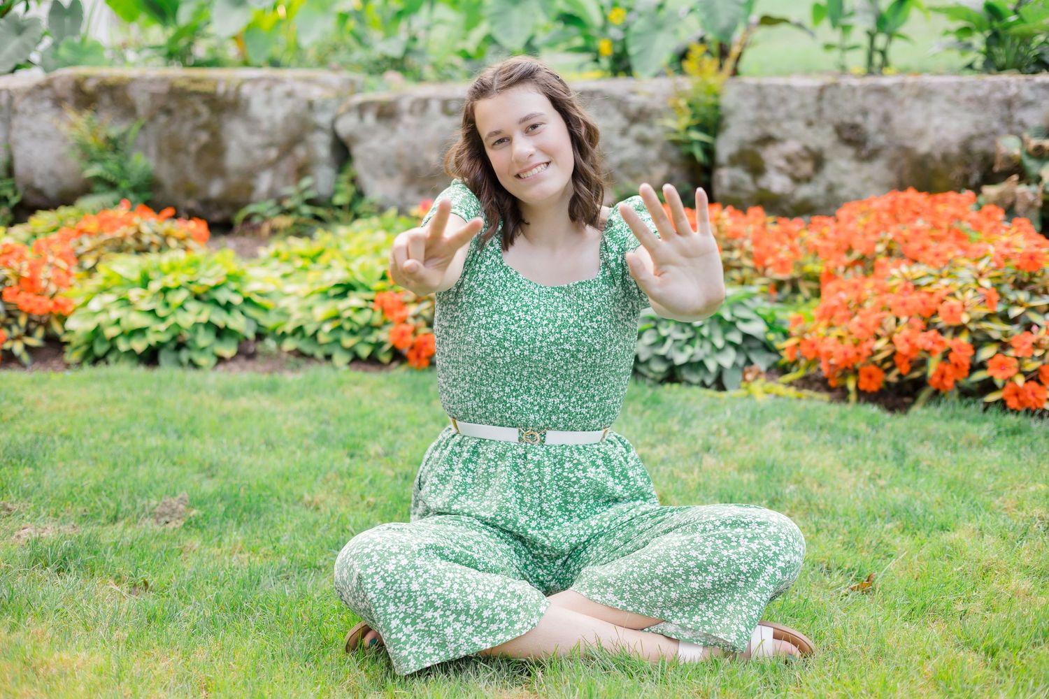 Woman in green floral jumpsuit sits cross-legged on grass near orange flowers.