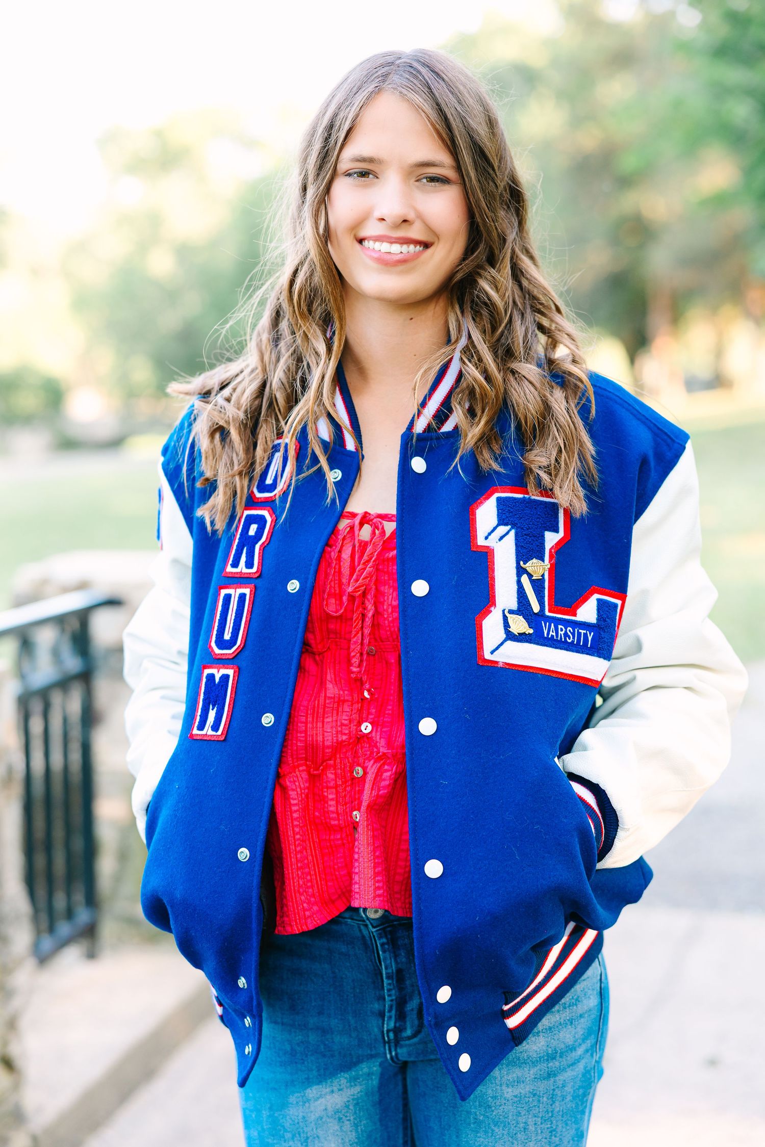Person wearing a blue and white varsity letterman jacket with red top and jeans standing outside on a sunny day.