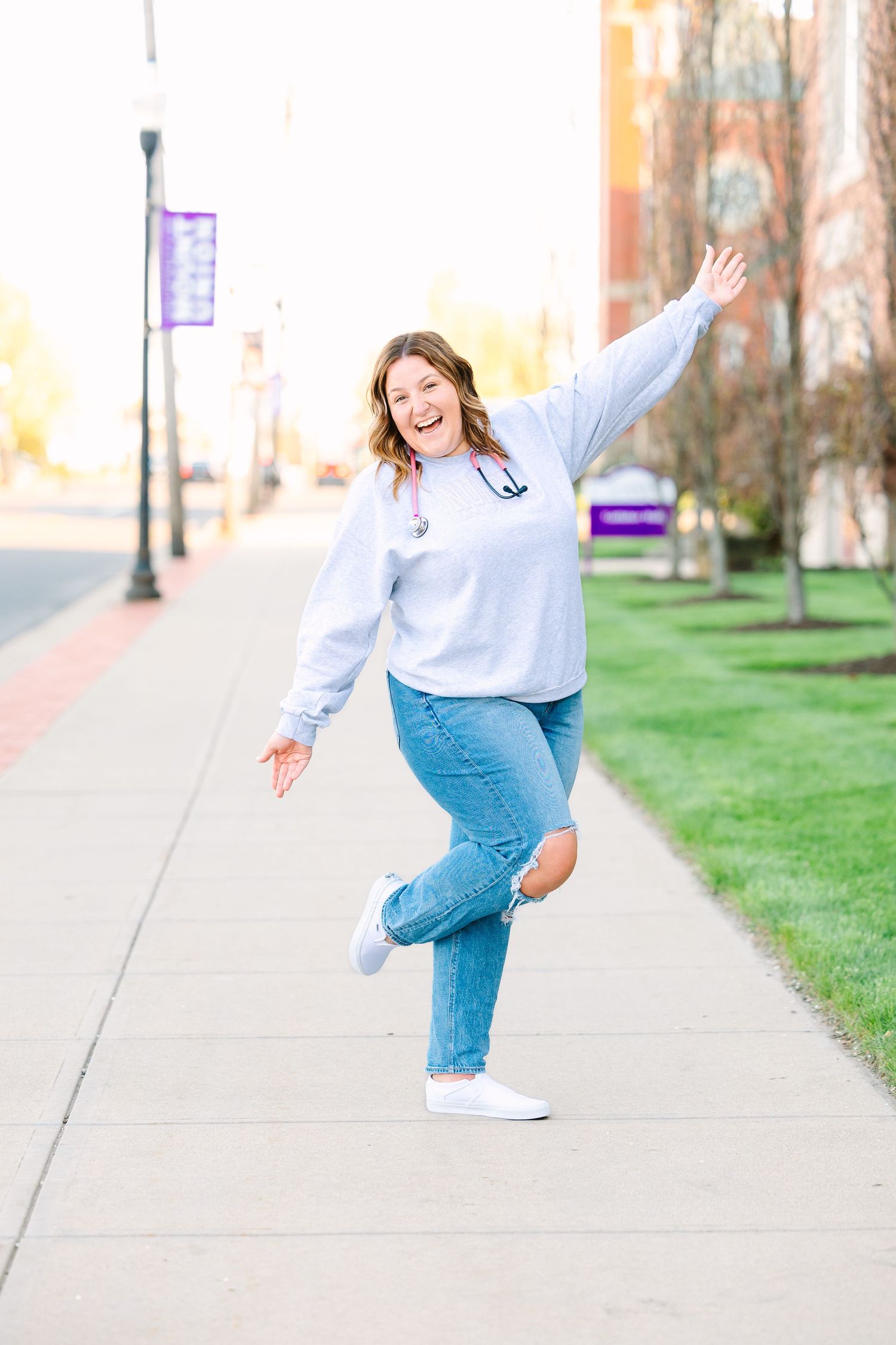 A person in a light blue sweater and jeans strikes a playful pose on a sidewalk next to a grassy area on a sunny day.