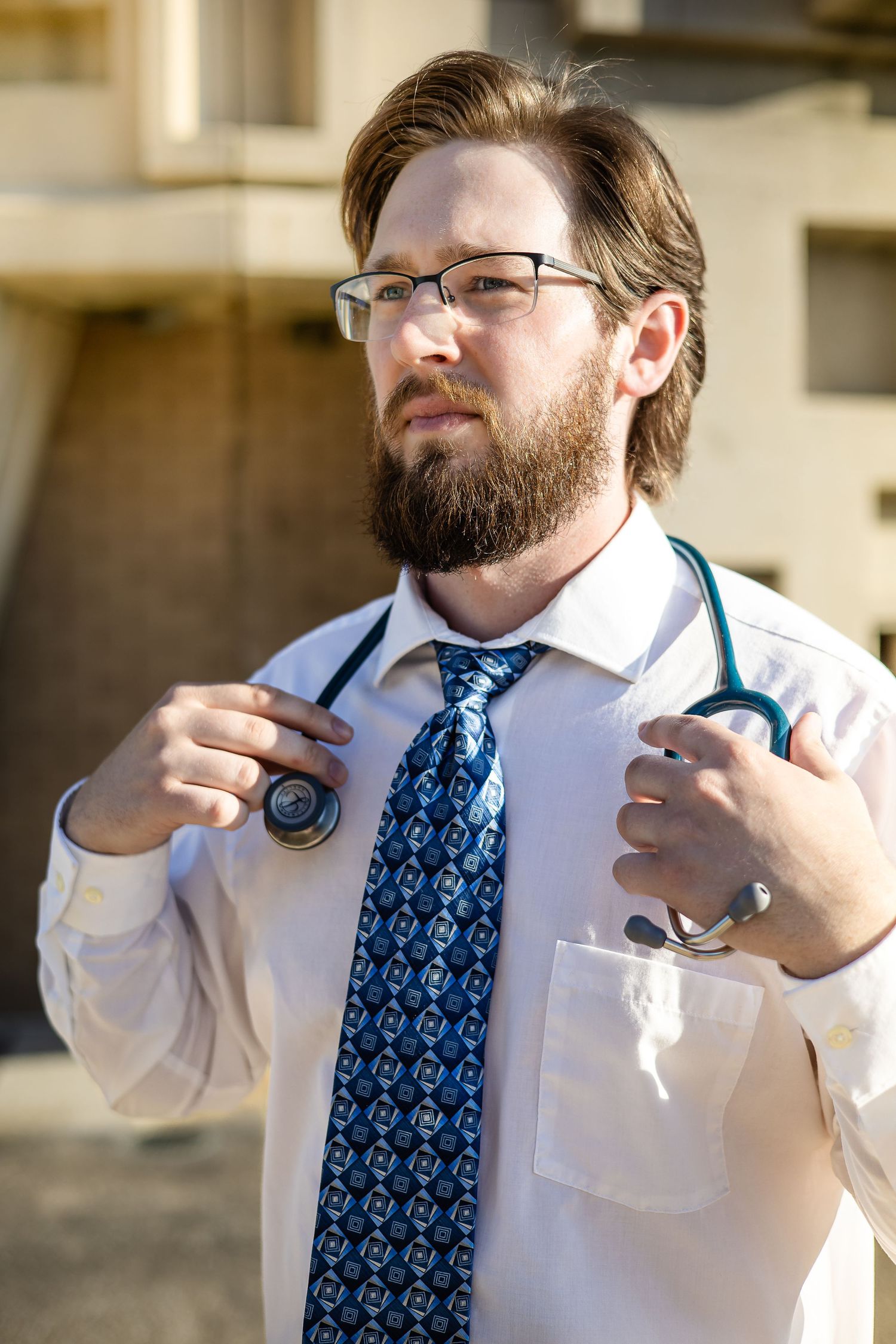 Medical professional adjusts stethoscope while wearing blue patterned tie and white coat.