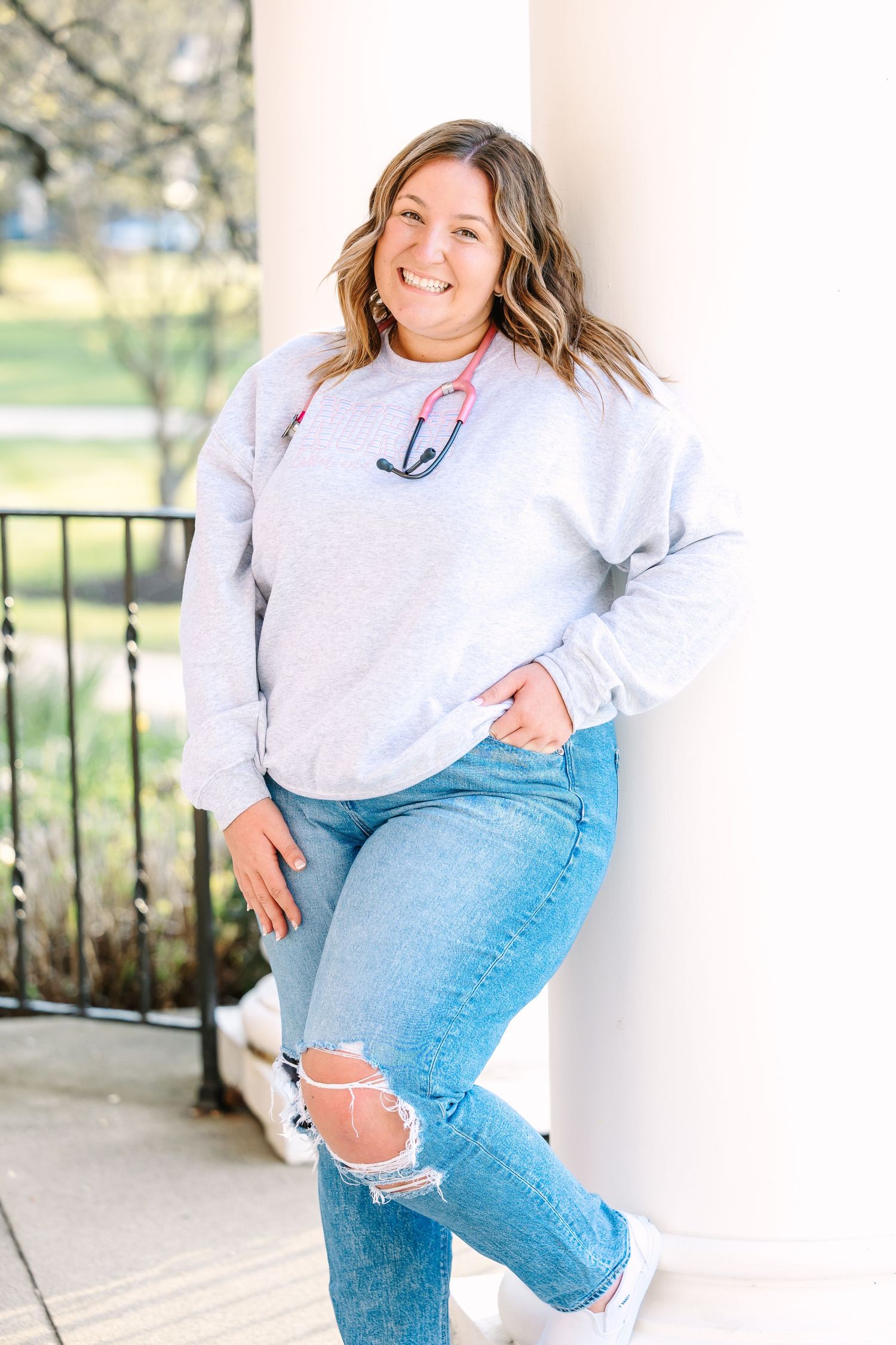 Person wearing grey anchor sweatshirt and ripped jeans poses casually against white column outdoors.