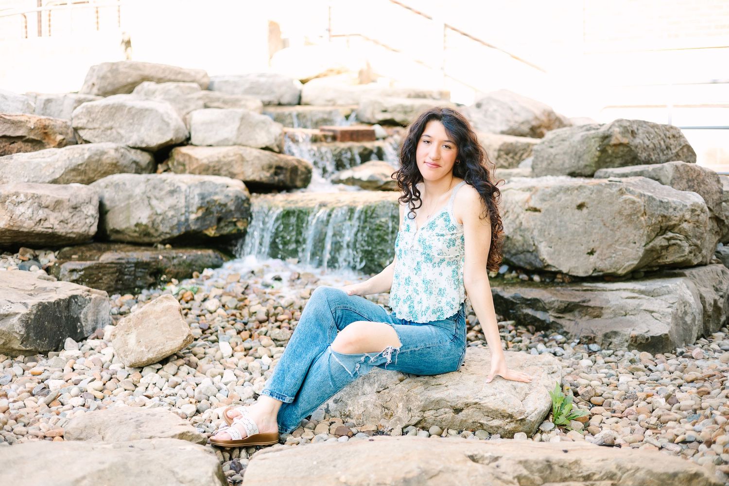 A person in a floral top and jeans sits casually on rocky terrain by a waterfall.