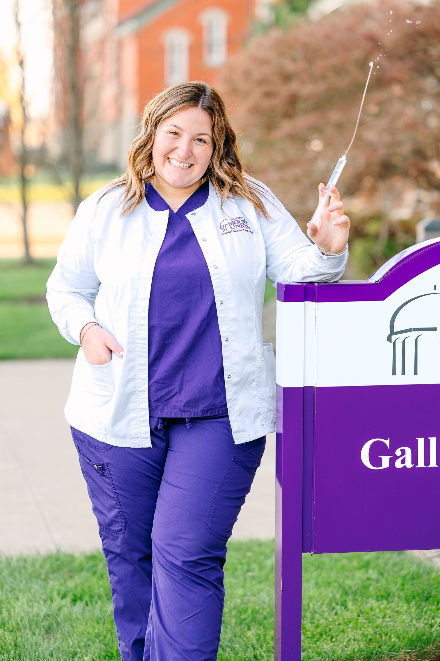 Healthcare professional in white coat and purple scrubs stands smiling by university sign on campus.