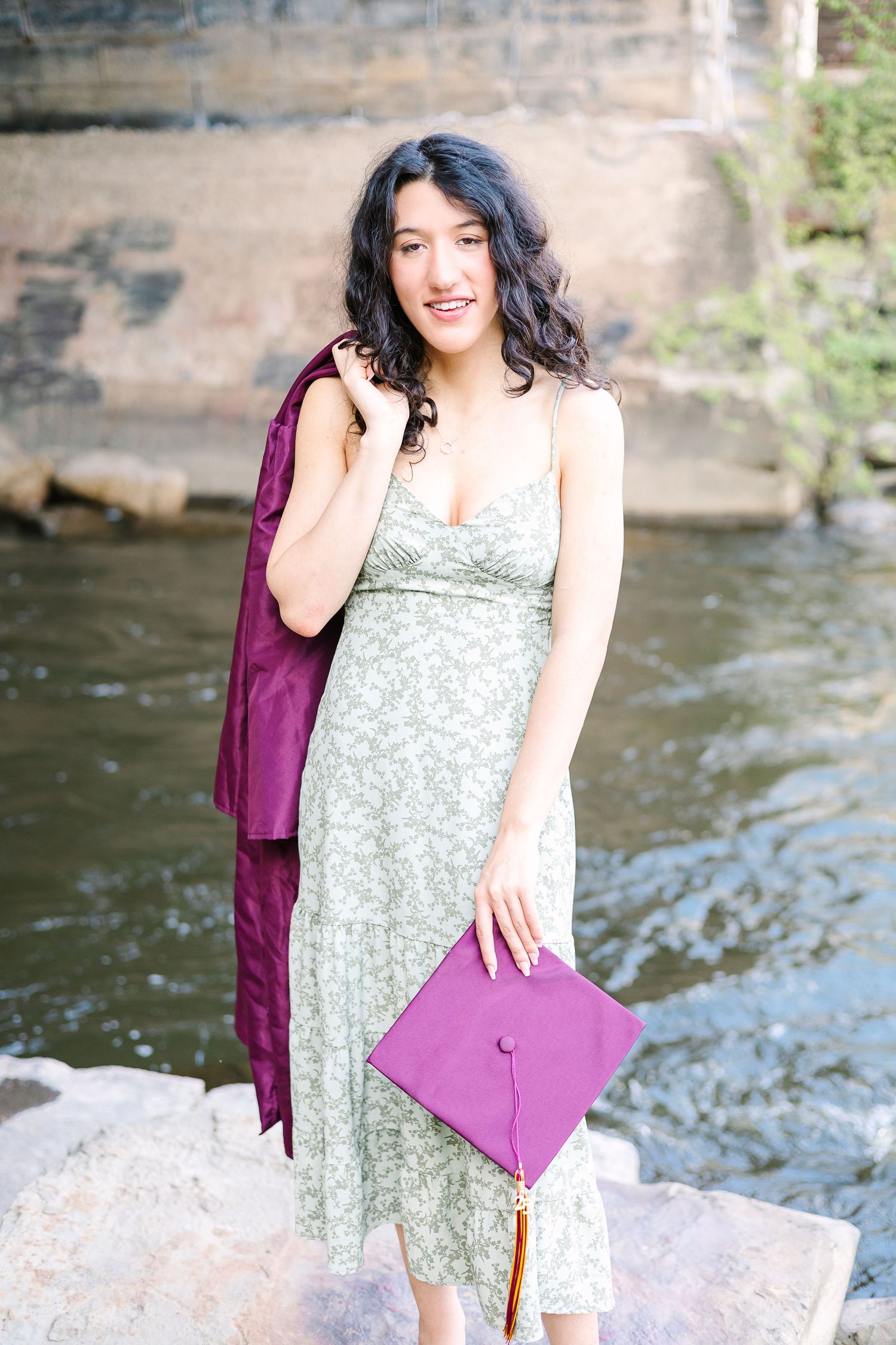A graduation photo featuring a green floral dress and burgundy cap and gown by a waterfall.