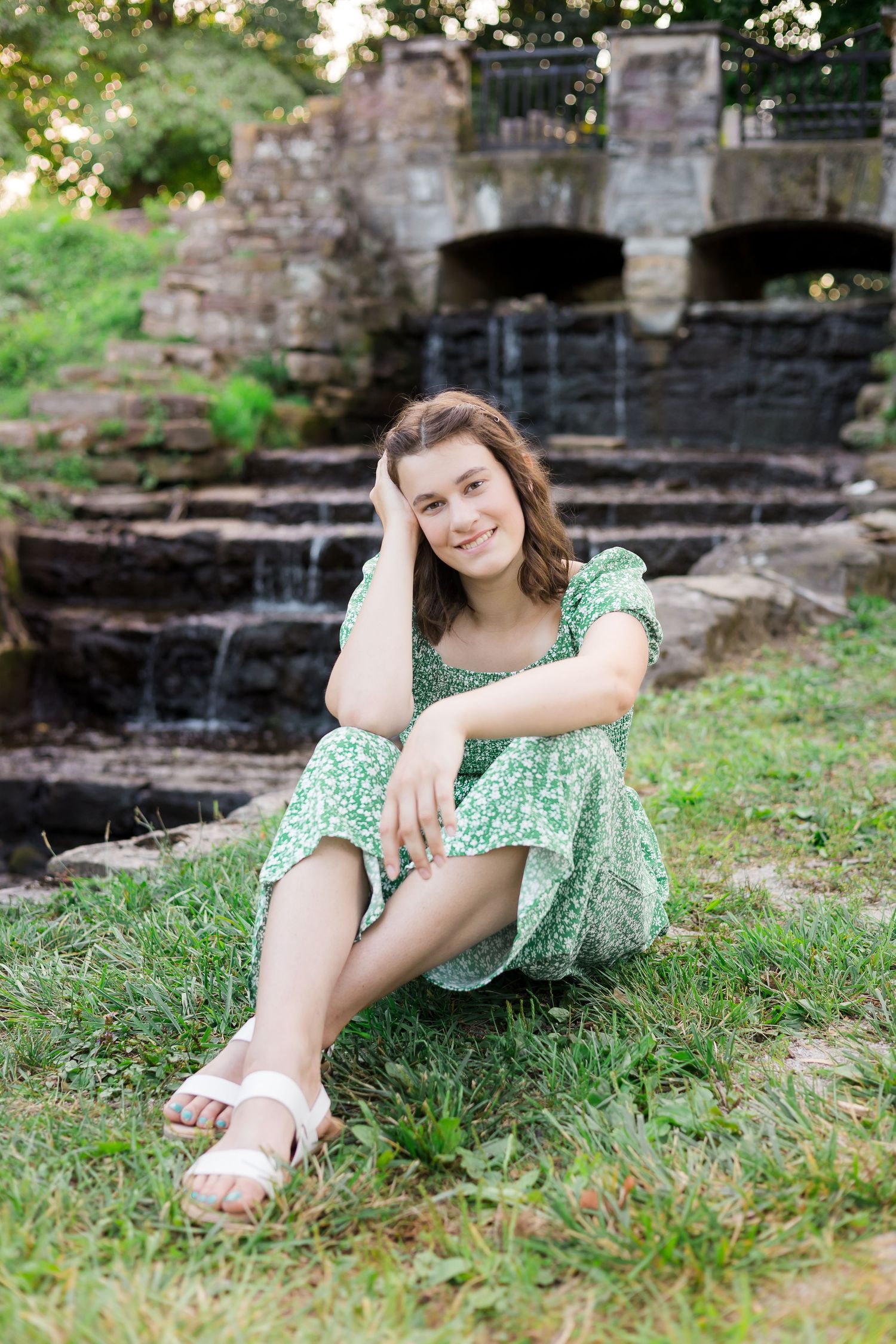 Woman in green dress relaxes by stone waterfall structure in garden setting.