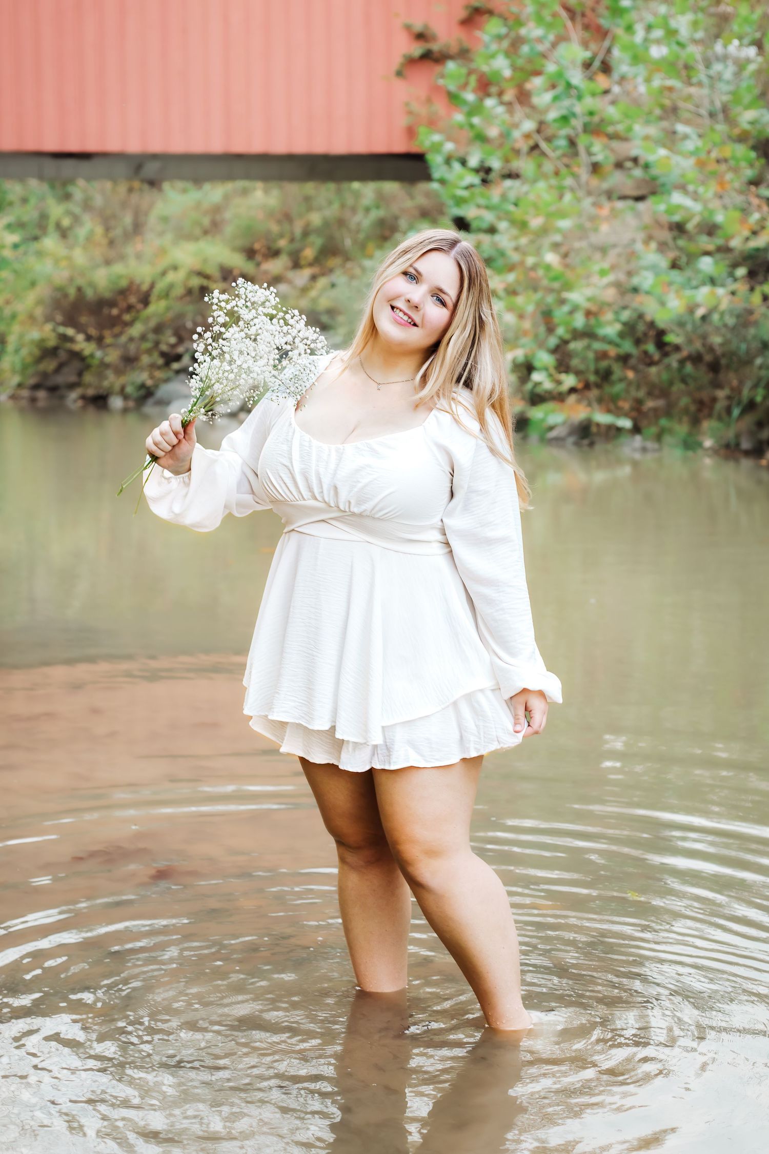 A person in a white dress stands in shallow water holding baby's breath flowers against a rustic red background.