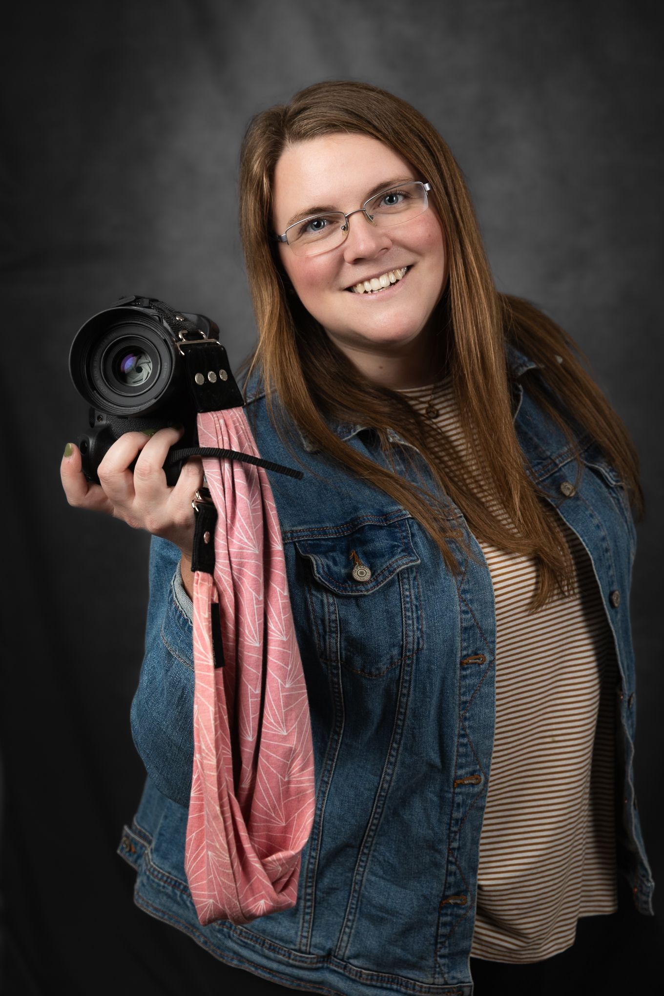 Professional photographer wearing glasses and denim jacket holds camera against dark studio background.