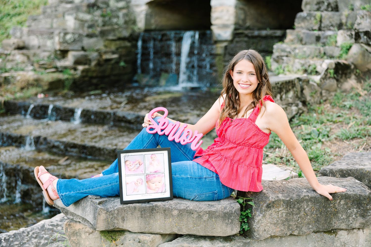 Woman in red top and jeans reclines on stone wall by cascading waterfall in outdoor portrait setting.
