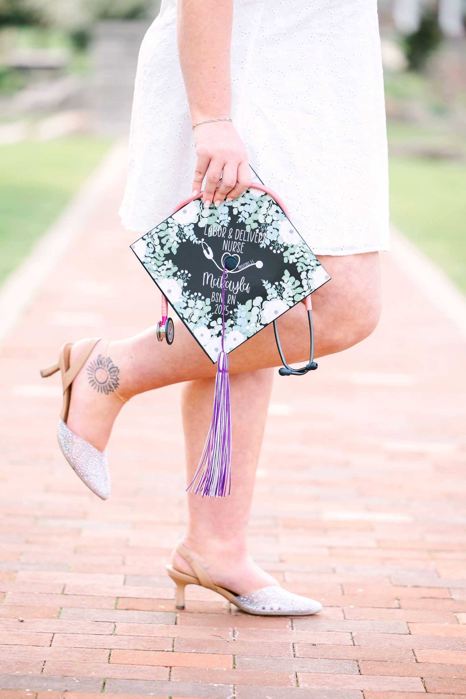 Graduate shows decorated mortarboard cap with purple tassel while wearing white dress and glittery shoes on brick path.
