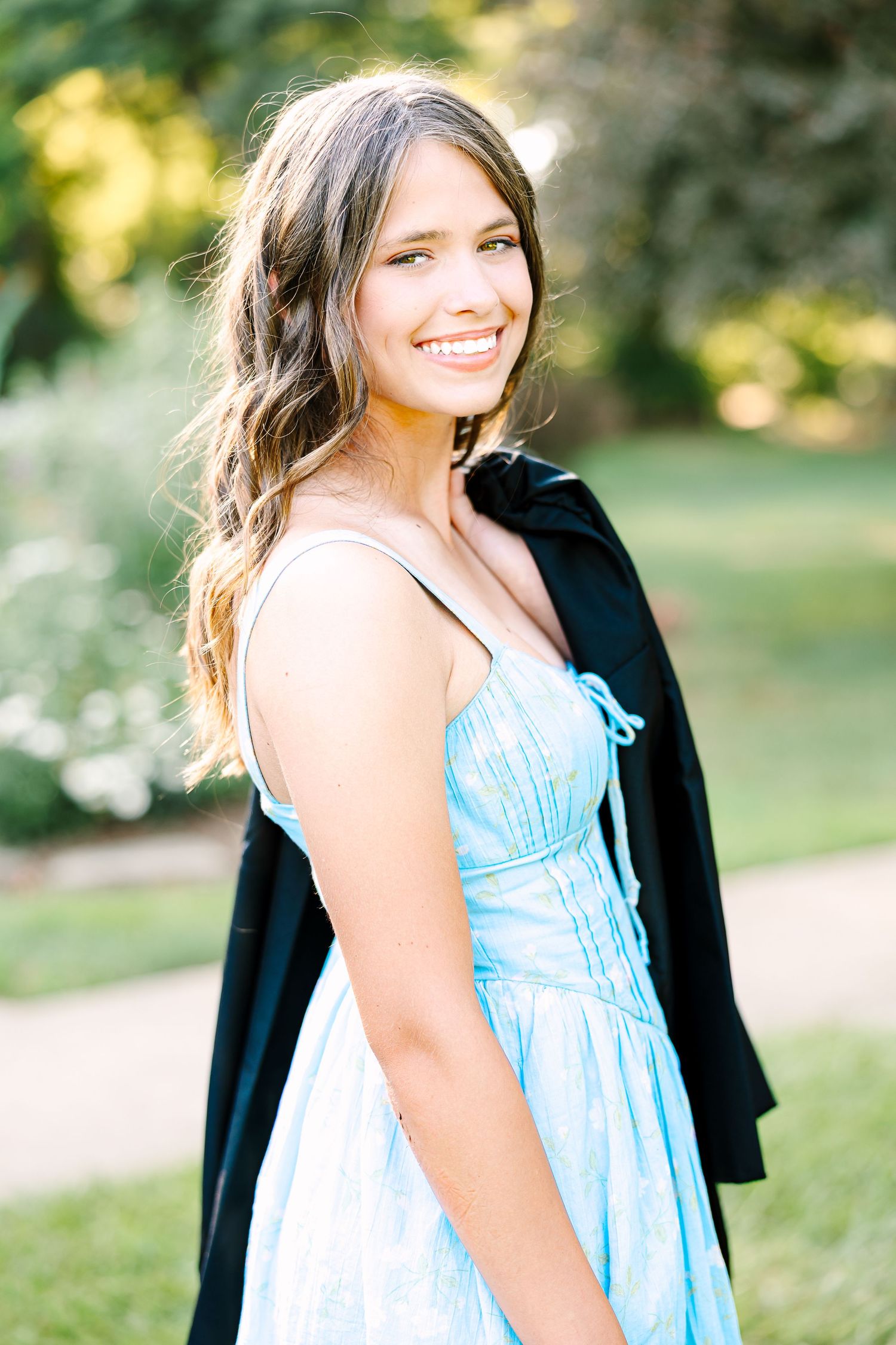 A person in a light blue dress and black jacket smiles while standing outdoors in a garden setting.