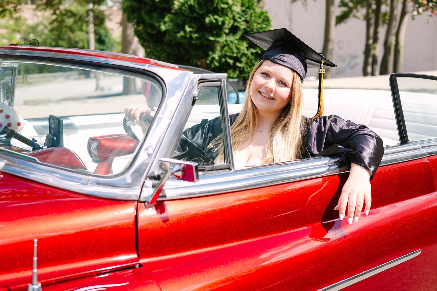 A graduate in cap and gown sits in a classic red convertible car on a sunny day.