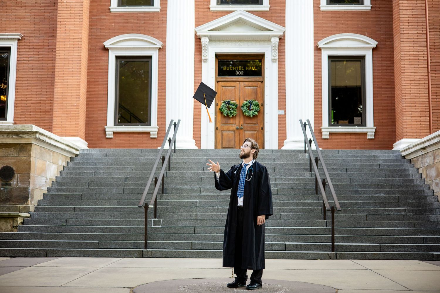 Graduate in academic regalia celebrates in front of grand brick building with white columns.