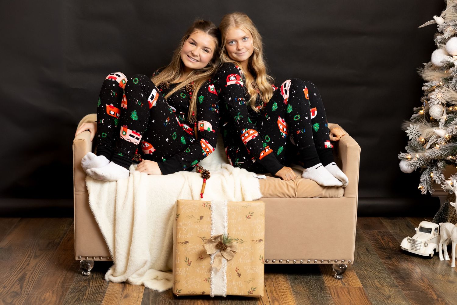Two friends in matching Christmas pajamas pose on a couch with wrapped presents and a decorated tree.