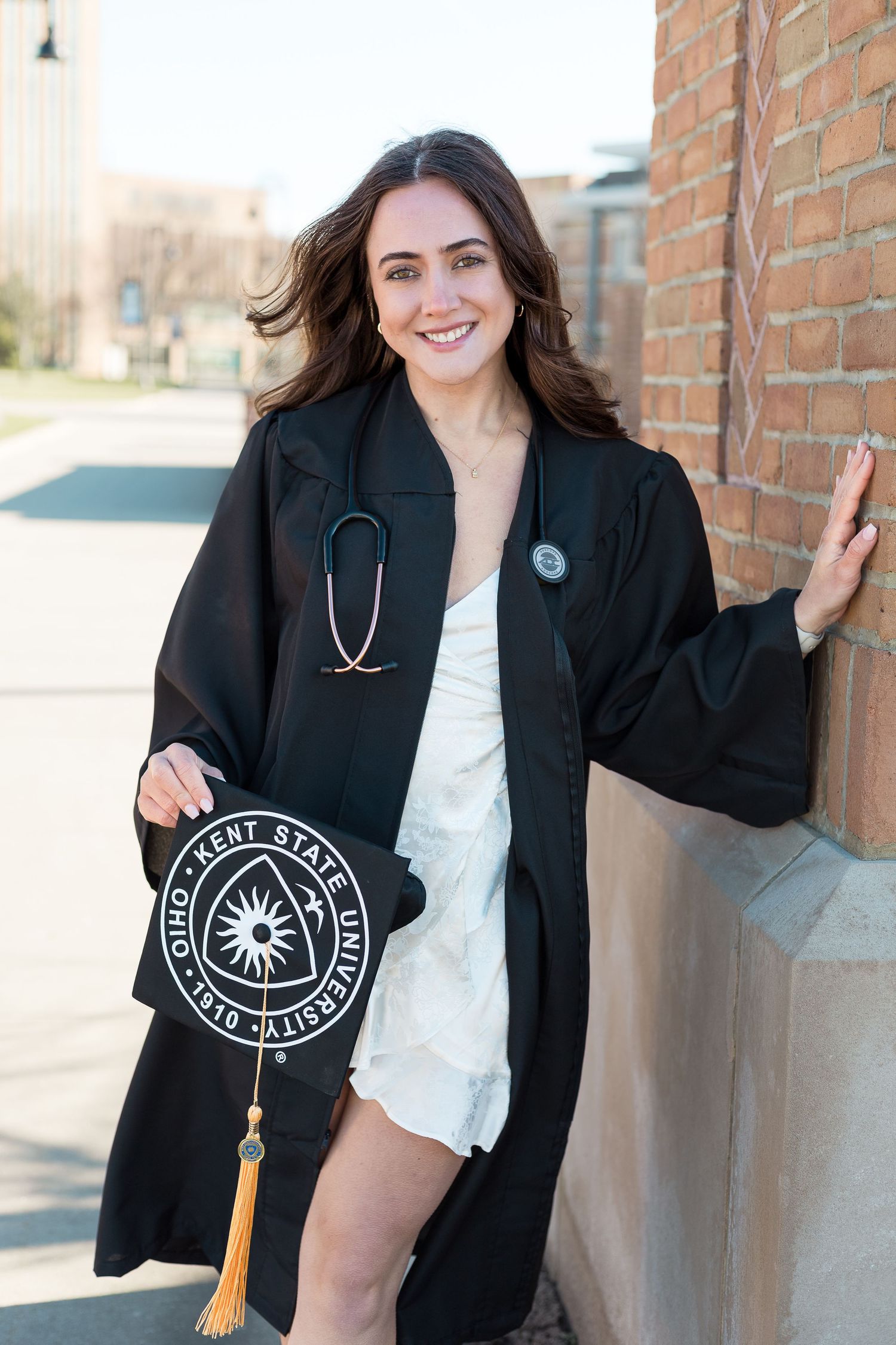 Medical graduate in black graduation gown poses with stethoscope and tassel outside brick building.