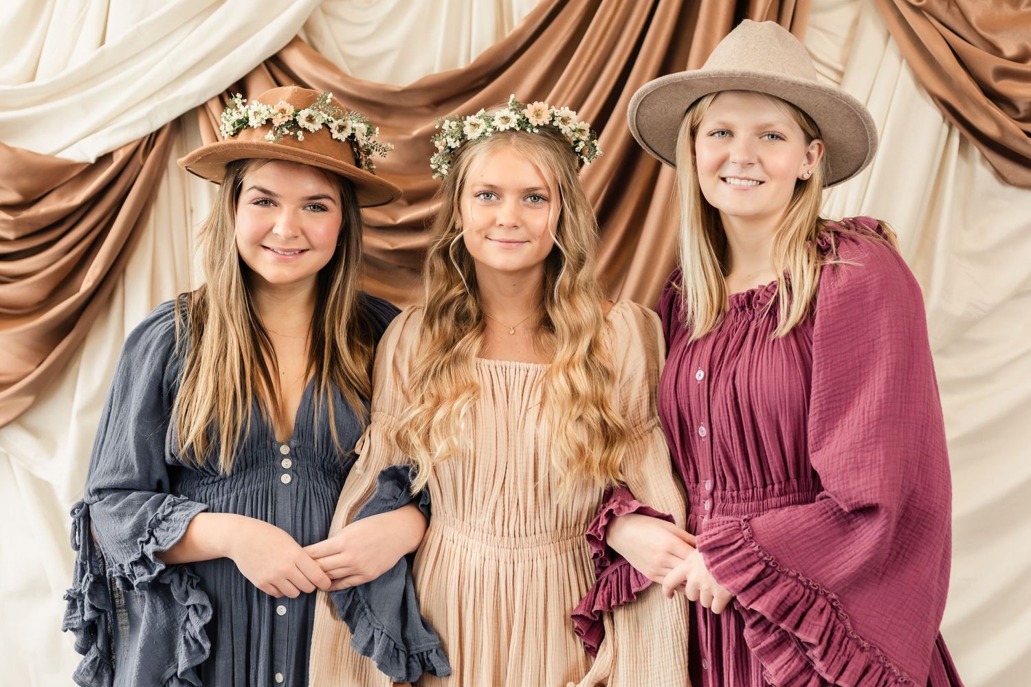 Three people in vintage-style dresses with floral crowns against draped fabric backdrop.