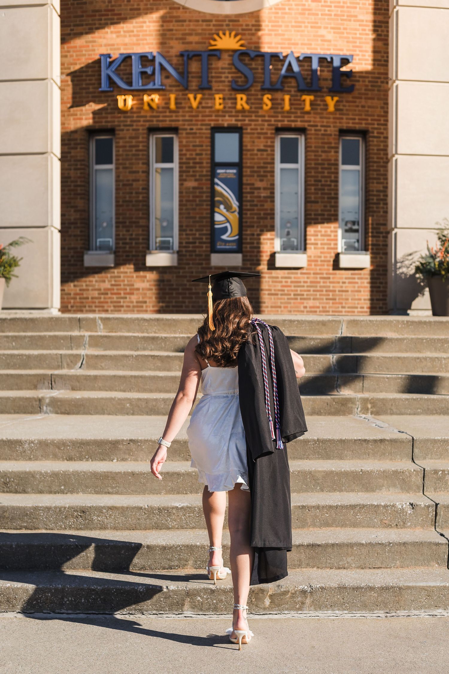 Graduate walking up stairs towards Kent State University entrance sign in cap and gown.