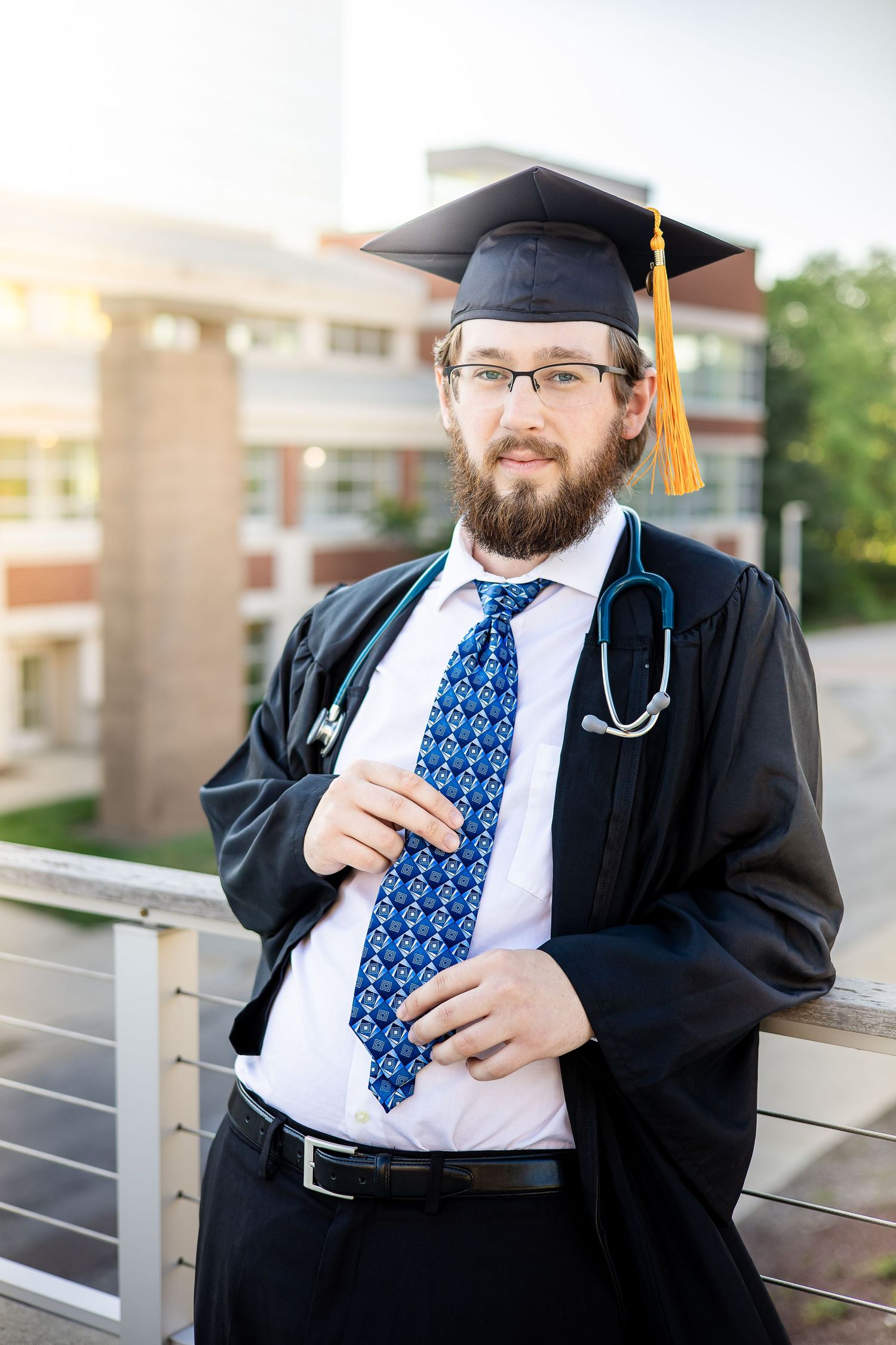 Graduate in academic regalia and medical attire poses outdoors with stethoscope.