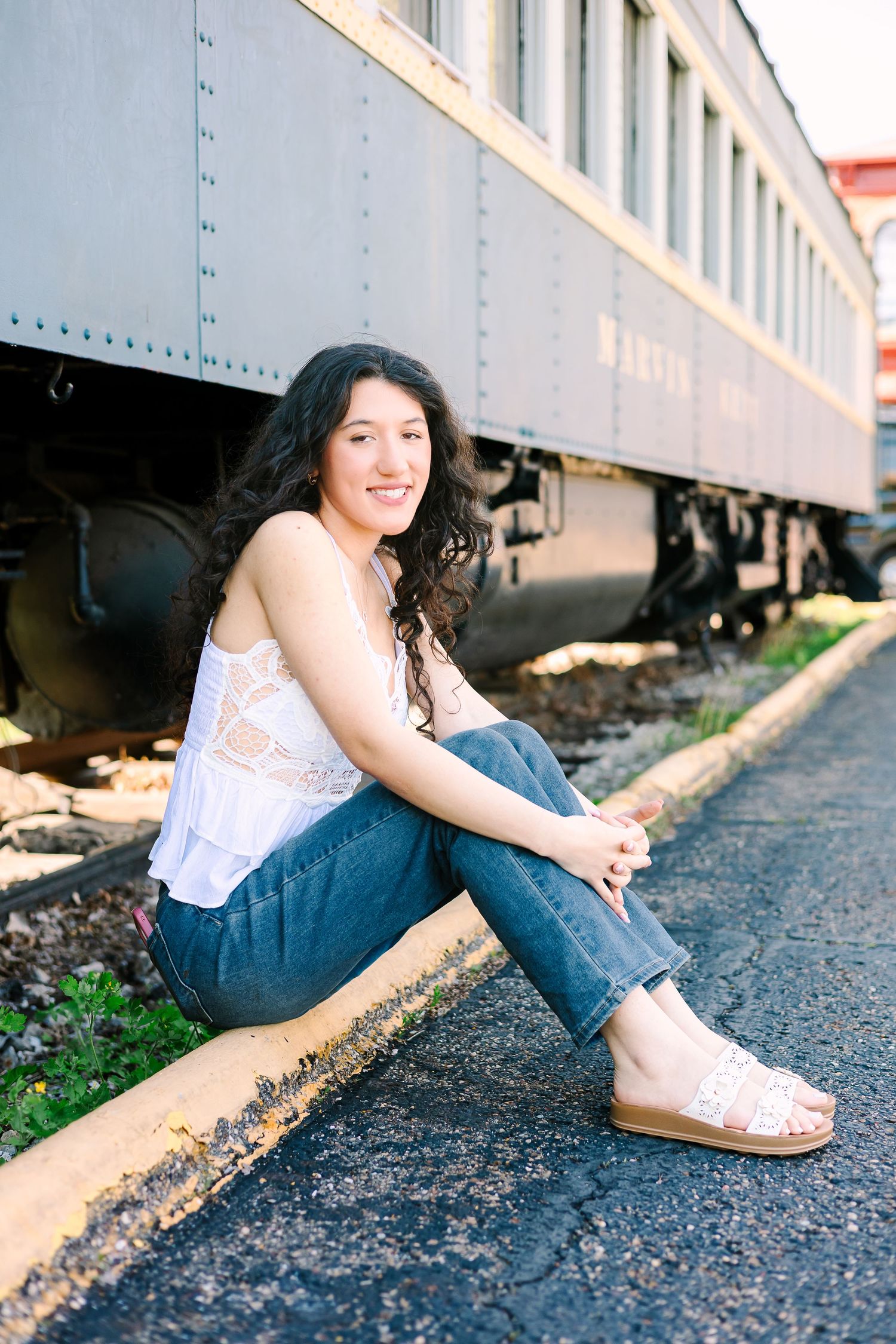 A person in a white top and jeans sits casually beside vintage train cars on the railroad tracks.