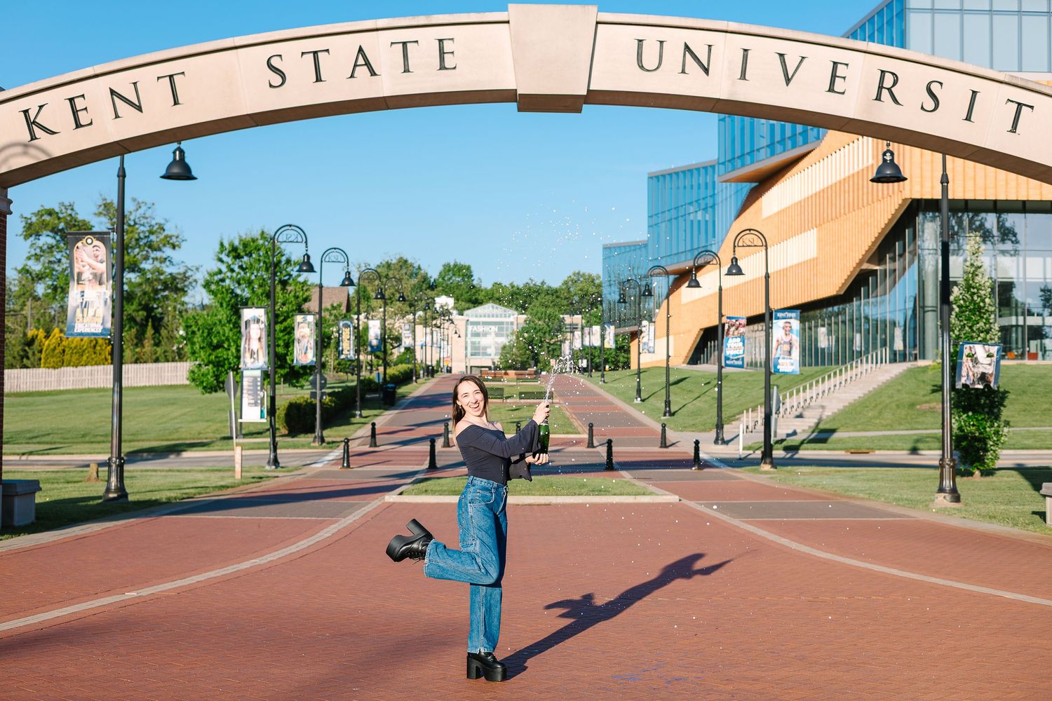 Student jumps joyfully under the Kent State University entrance arch on campus walkway during sunny day.