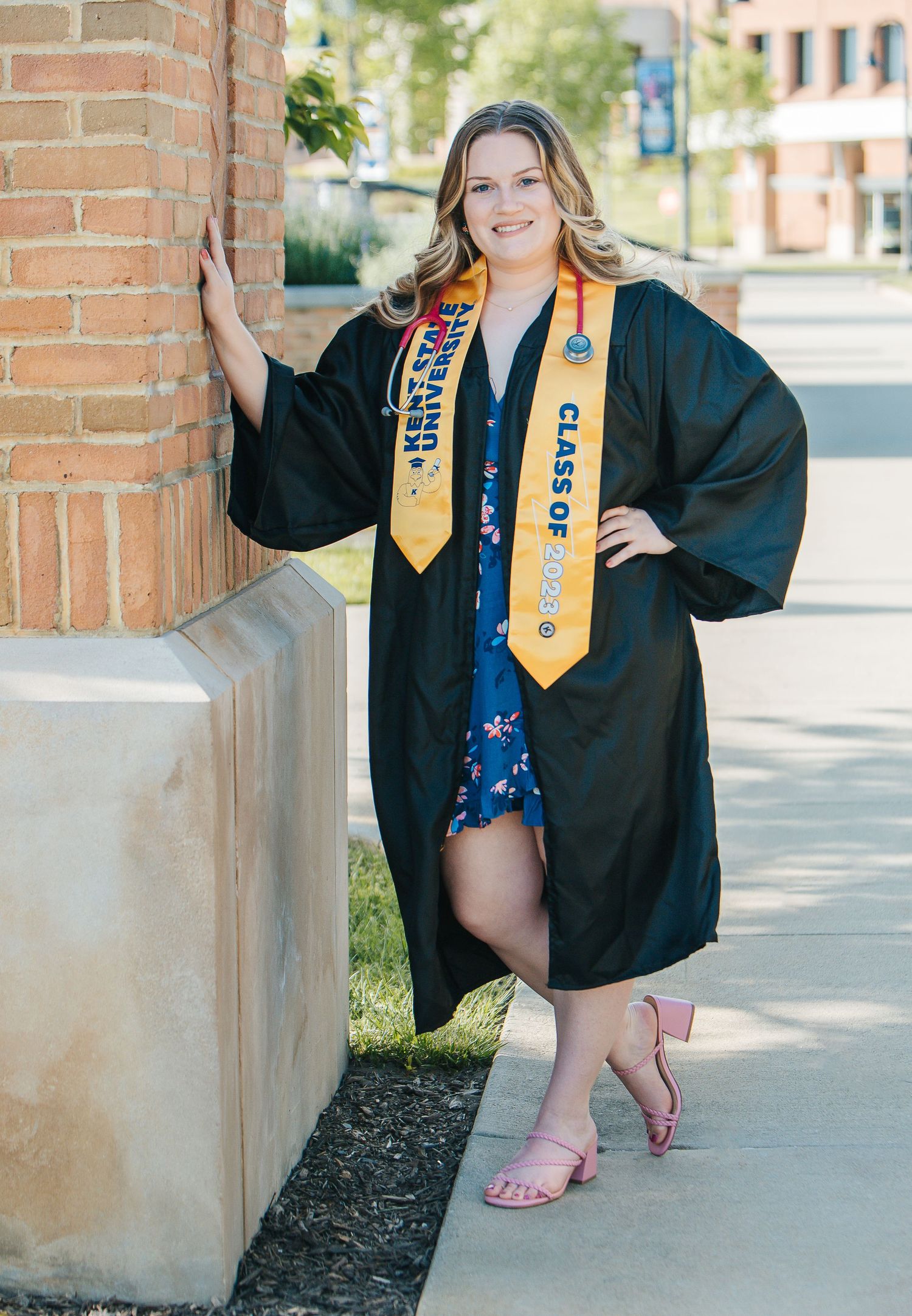 Graduate wearing academic regalia and gold stole poses confidently by brick wall.