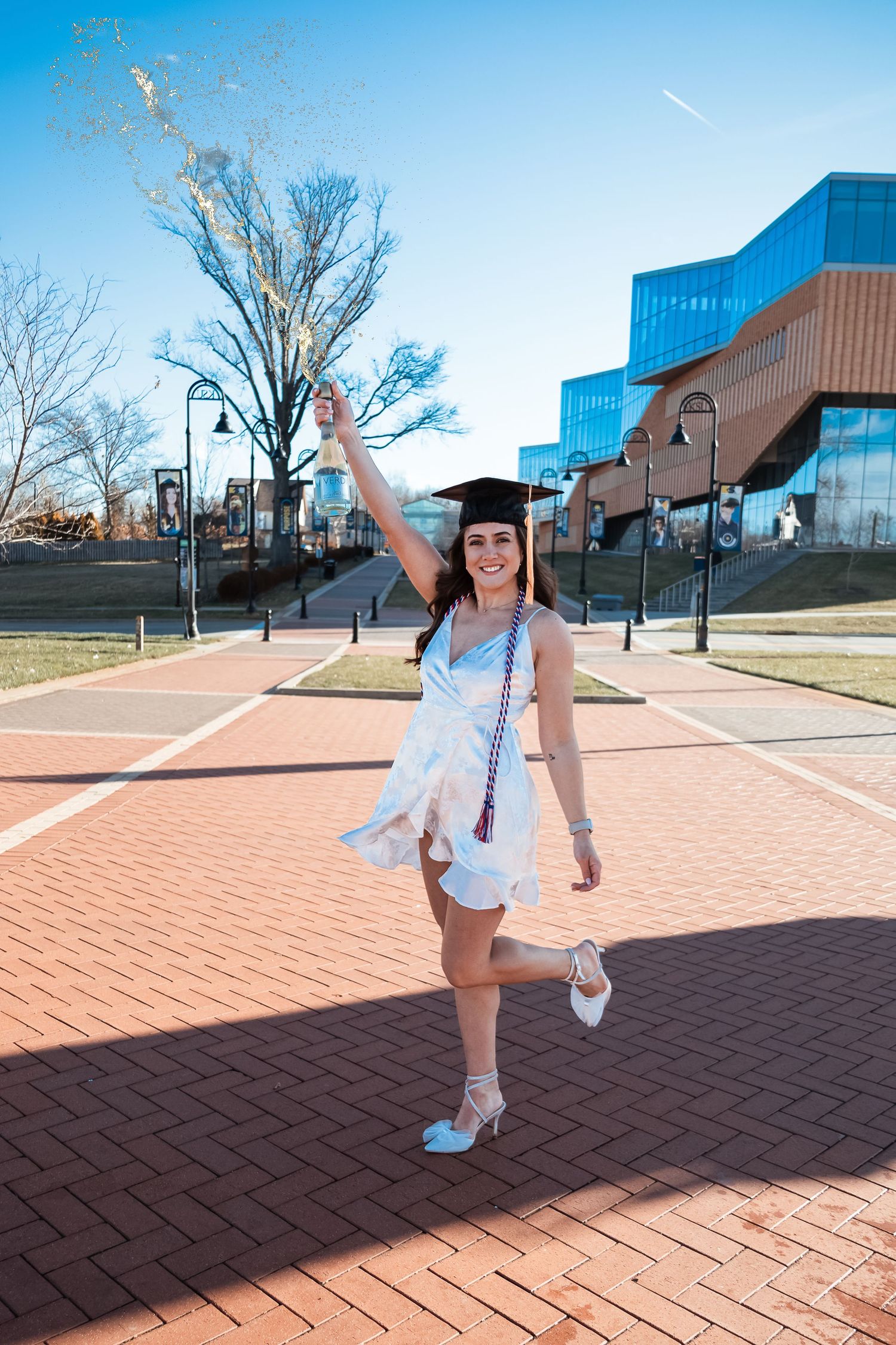 Graduate celebrating in white dress and cap in front of modern university building.