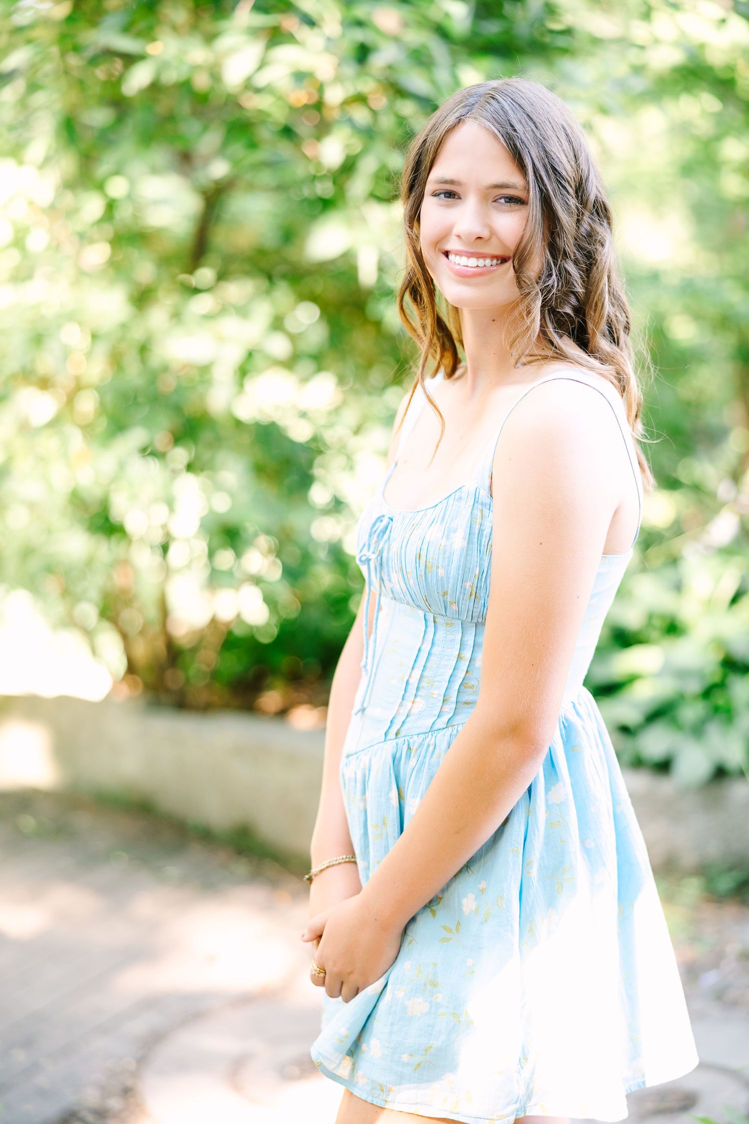 Young woman in light blue summer dress poses outdoors with greenery in background.