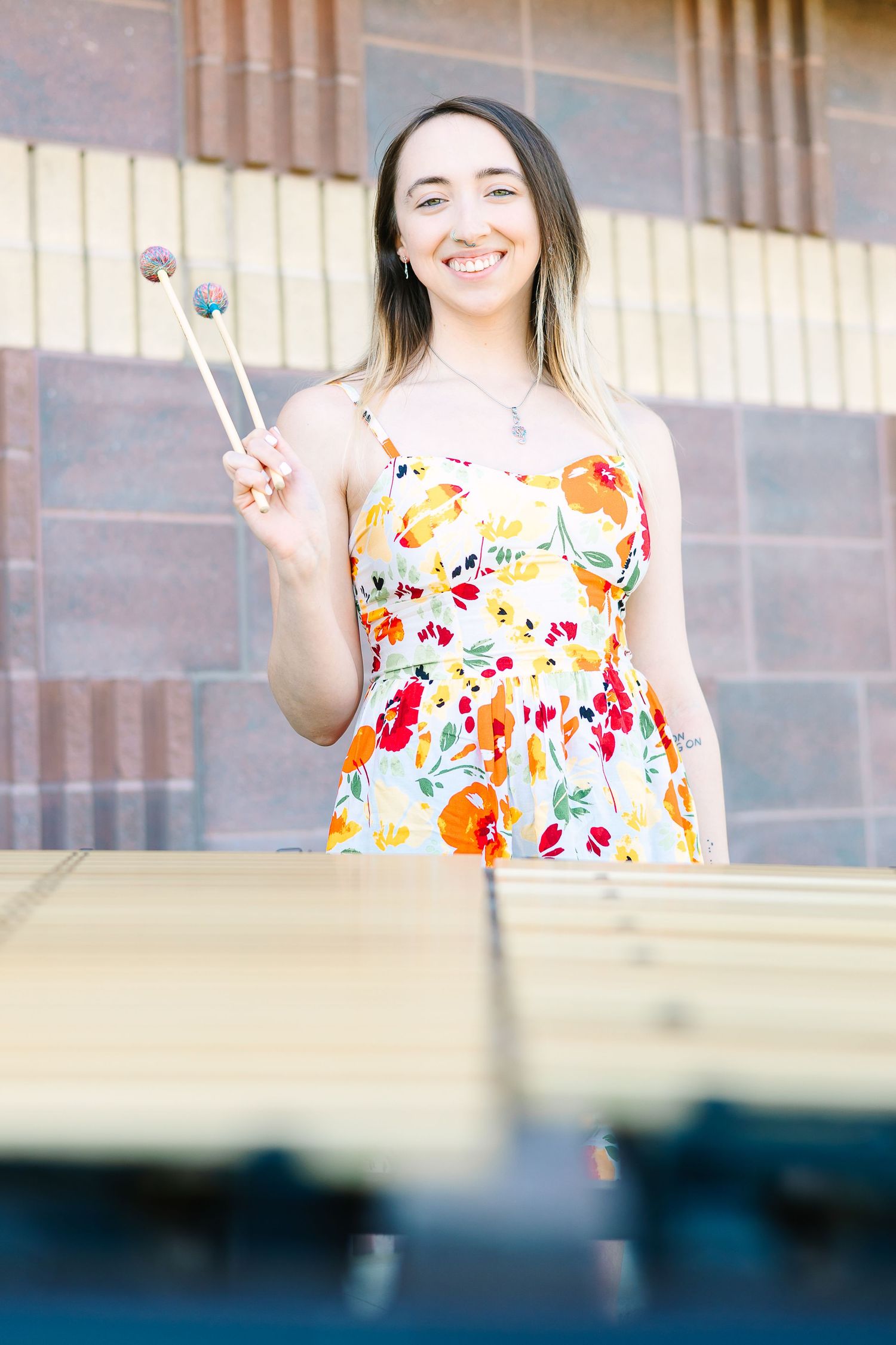 A percussionist in a bright floral sundress holding mallets stands behind a marimba instrument outdoors.