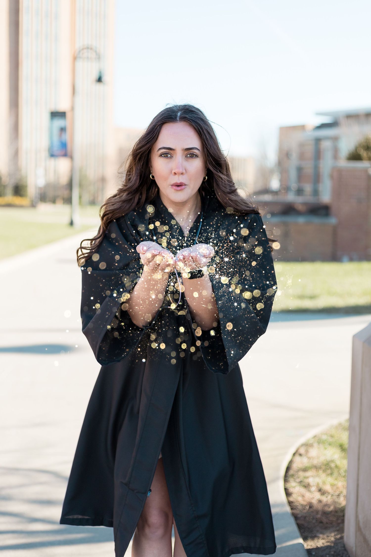 Graduate in black gown blowing glitter during outdoor graduation photoshoot.