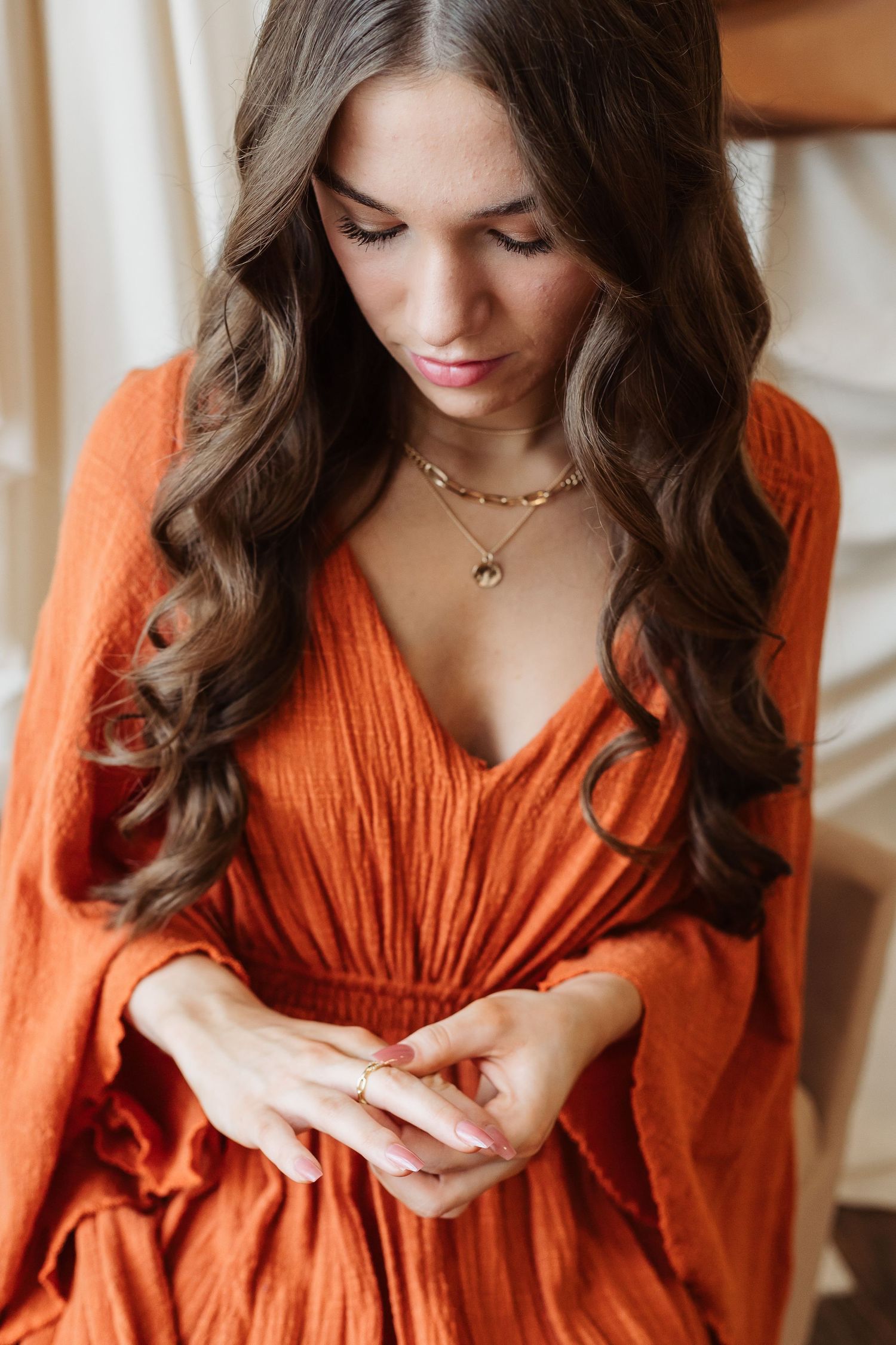 Close-up portrait in orange dress with delicate necklaces against neutral backdrop.