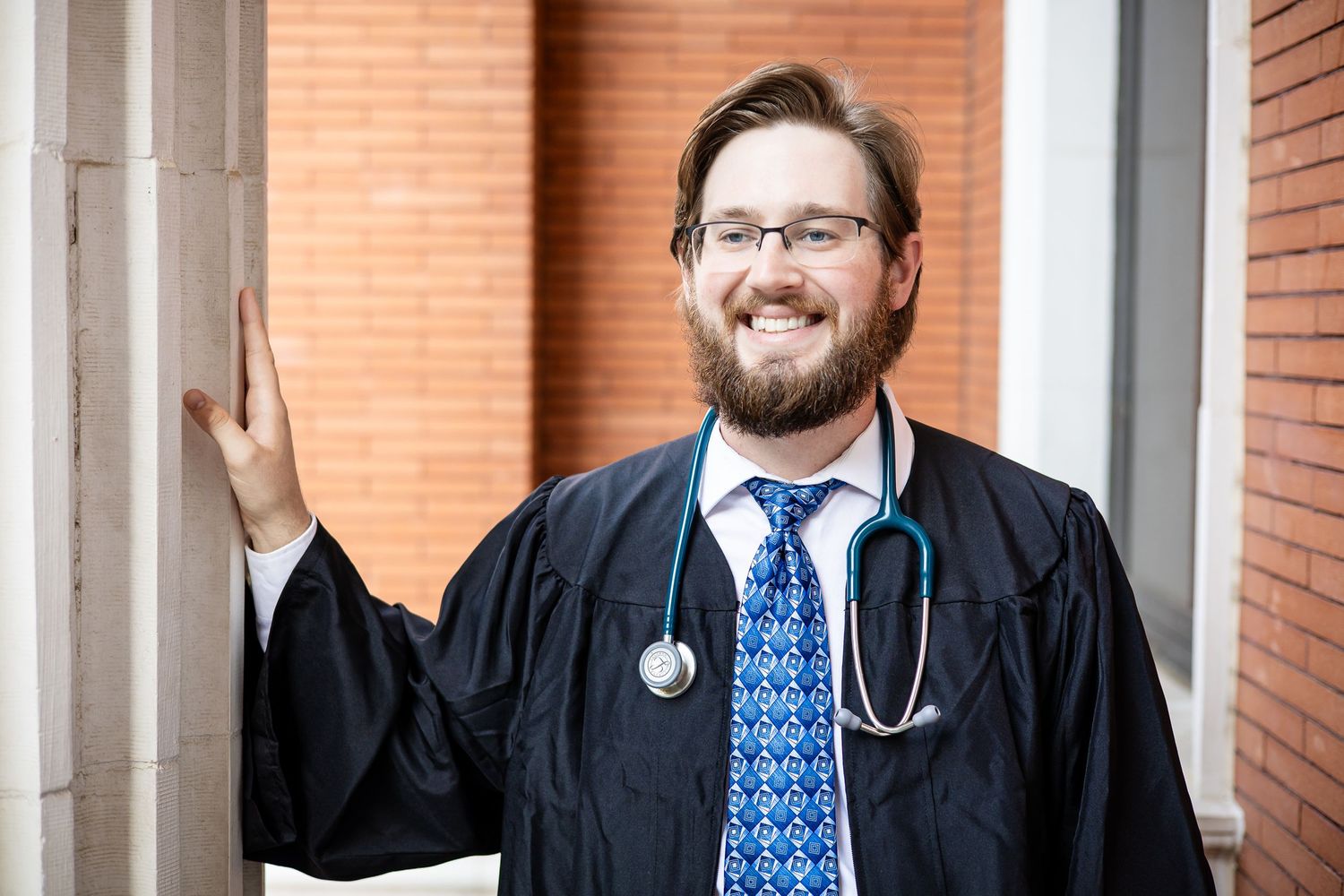 Medical graduate in academic regalia and stethoscope smiles near brick building.