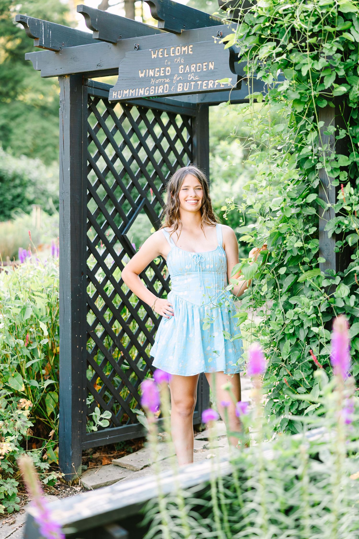 Woman in sundress stands in garden by lattice welcome sign surrounded by purple flowers and greenery.