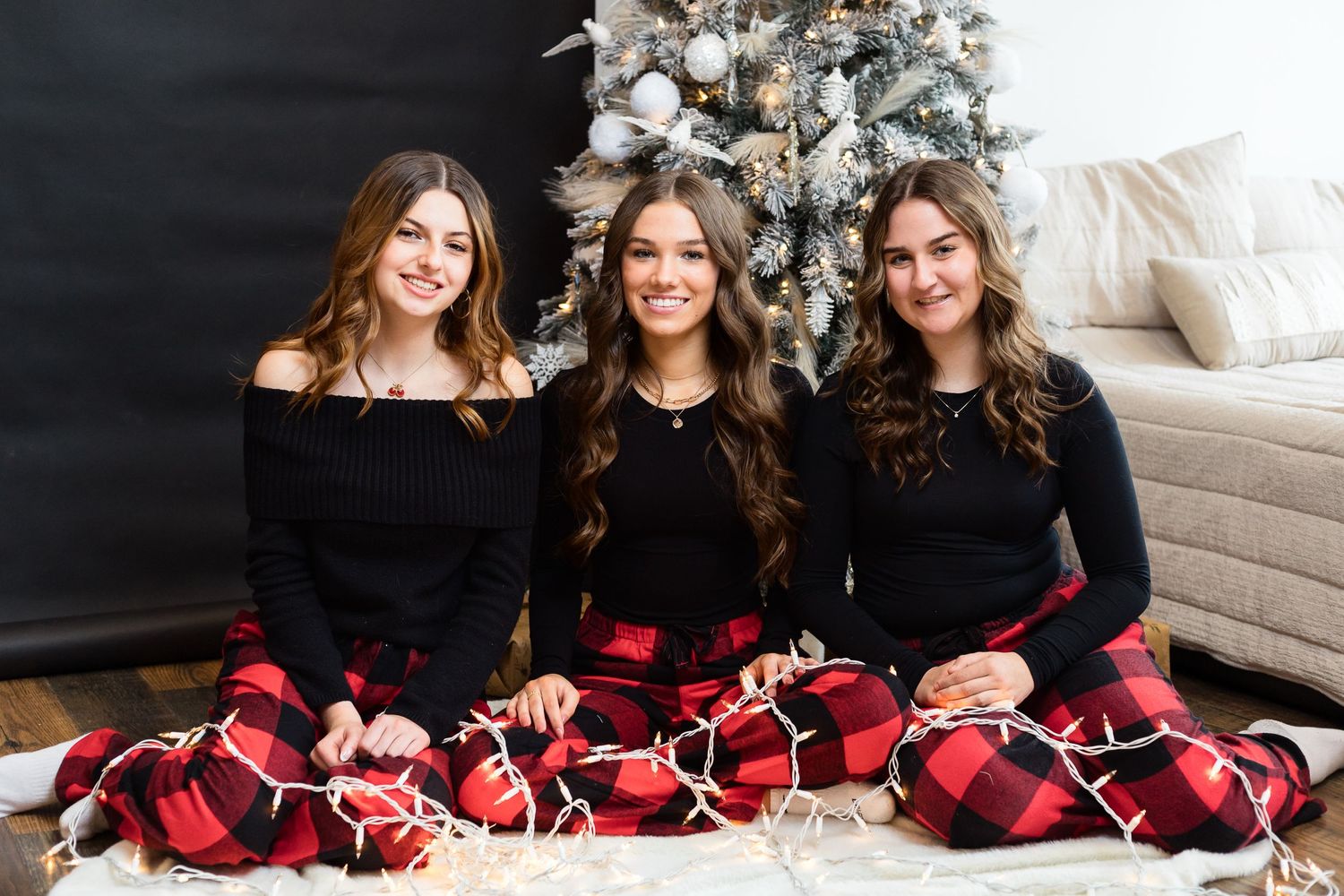 Three people wearing matching black tops and buffalo plaid pajama pants seated by Christmas tree.