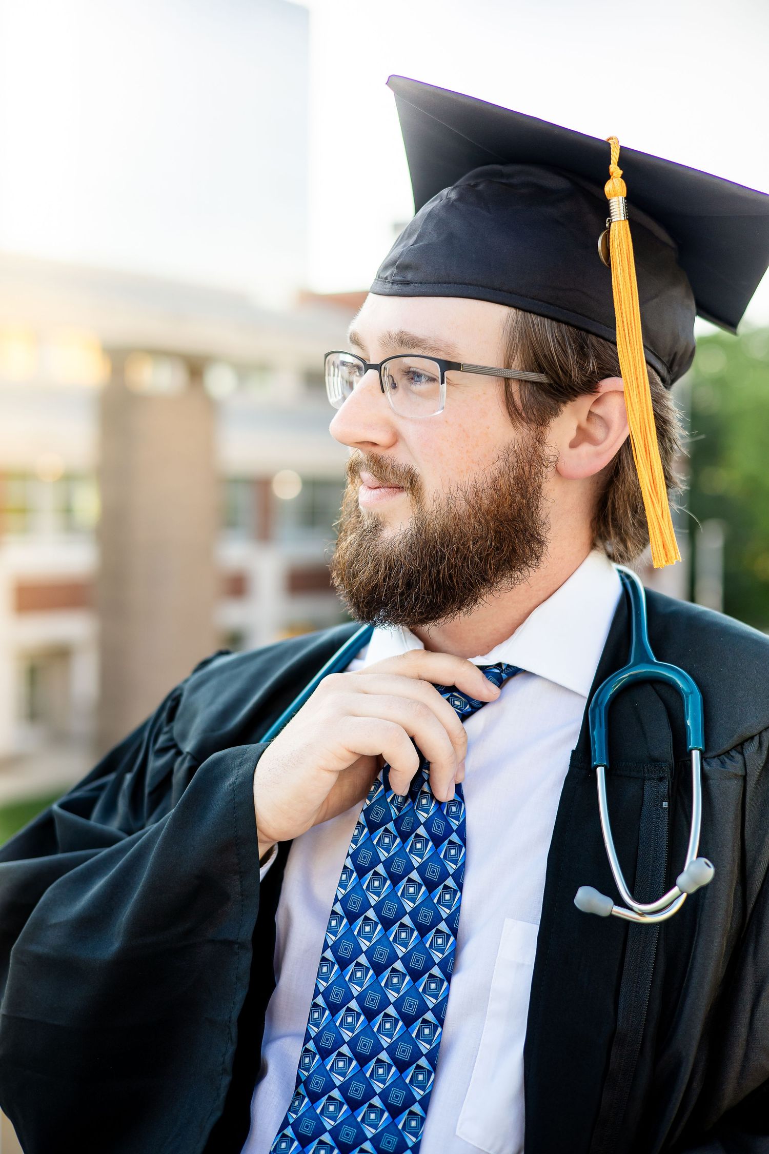 Medical graduate wearing cap and gown with stethoscope adjusts blue patterned tie.