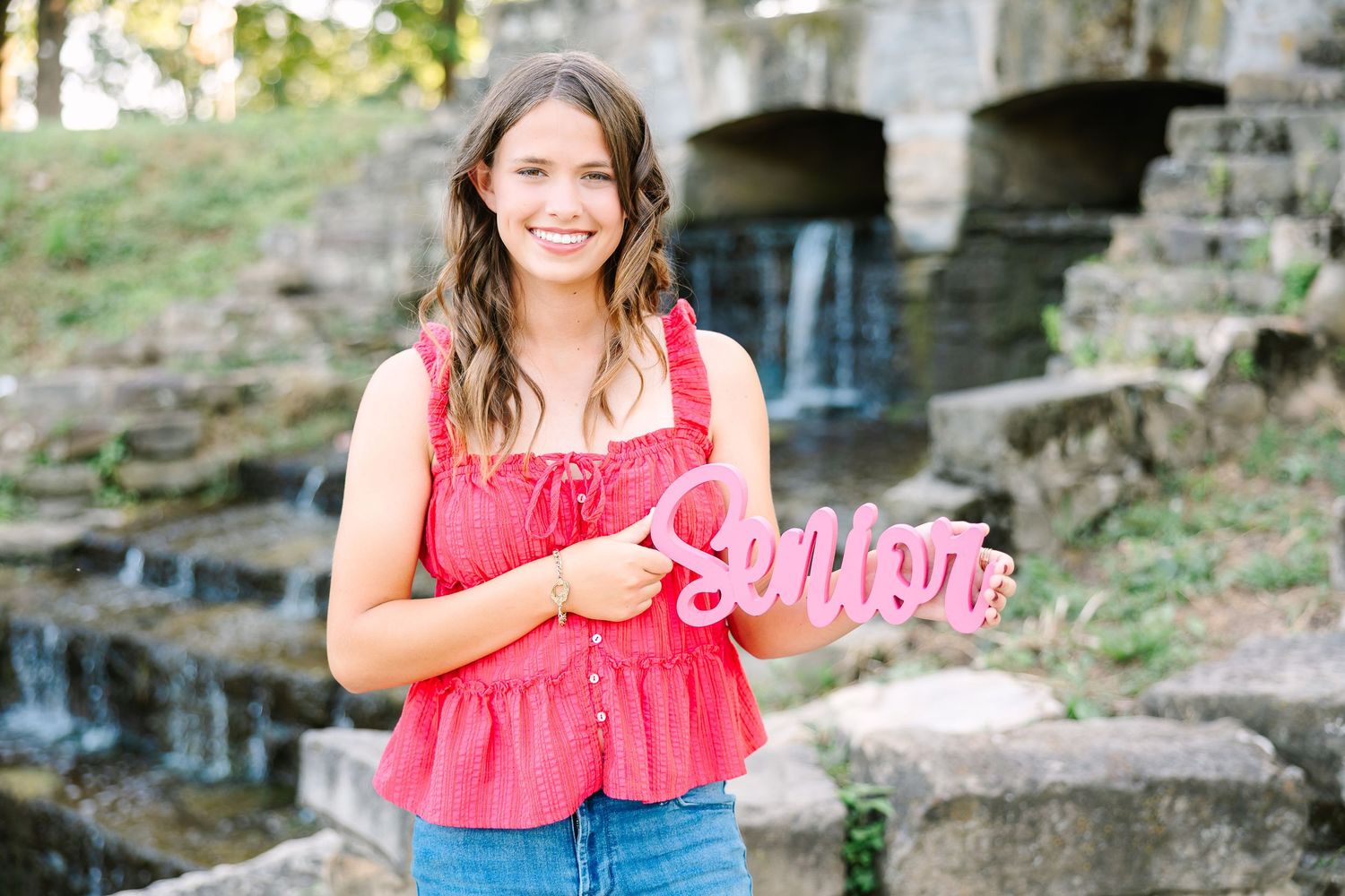 Someone in a red top and jeans stands outdoors holding a pink sign by a stone wall with arches.
