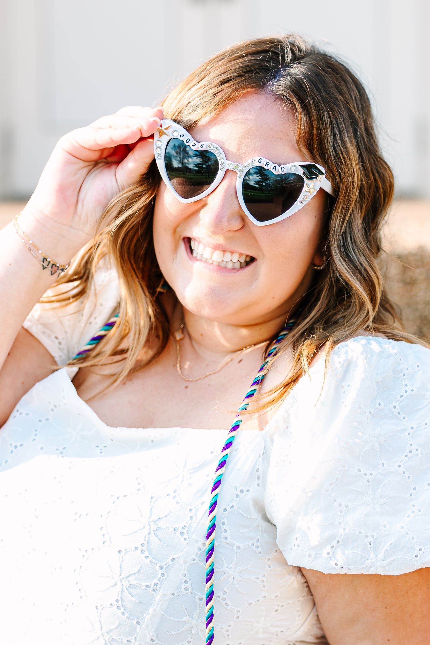 Someone in a white eyelet dress and heart-shaped sunglasses smiles while touching their frames in a sunny outdoor setting.