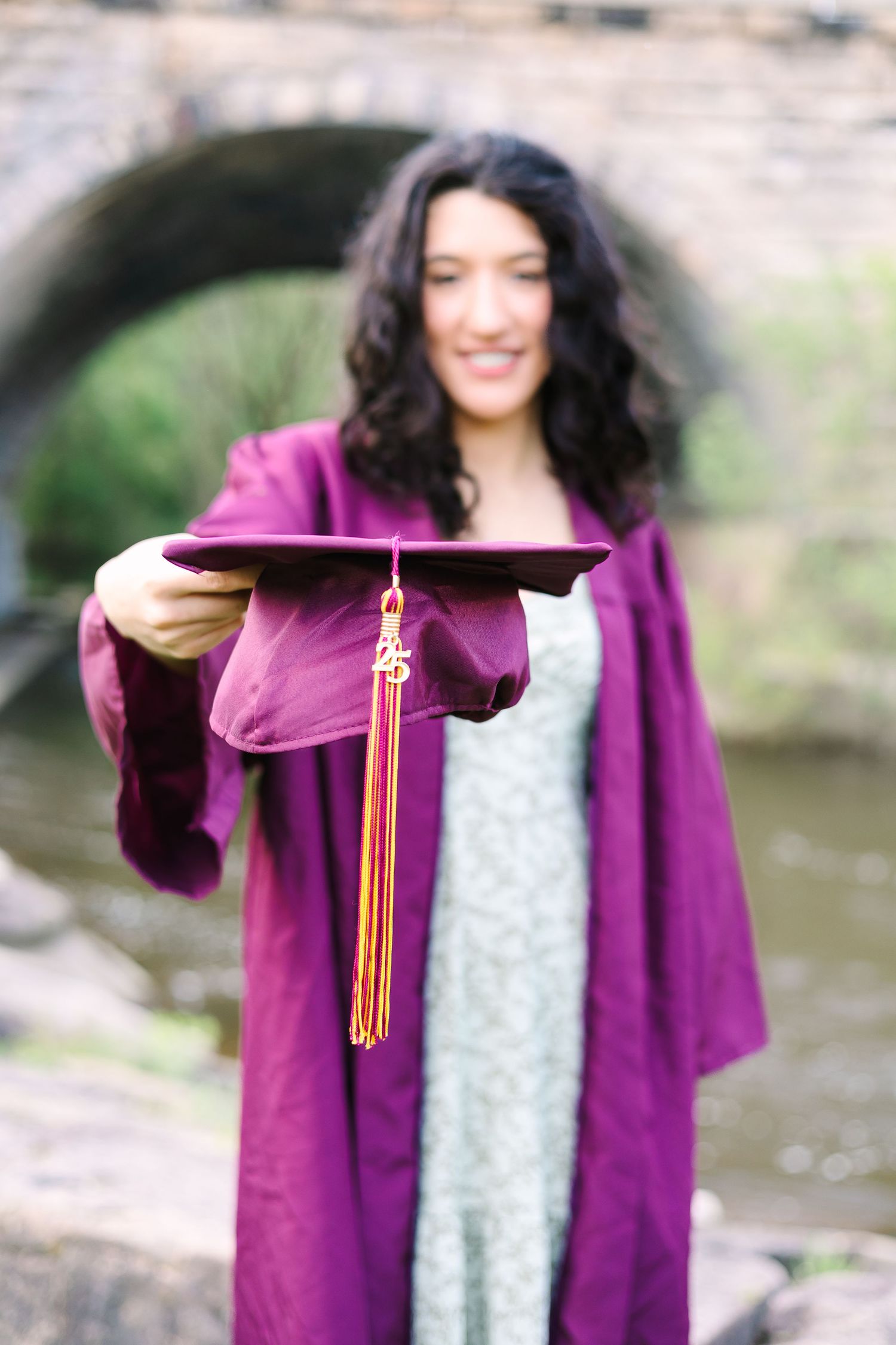 Graduate in burgundy cap and gown holds graduation cap with gold tassel outdoors near stone bridge.