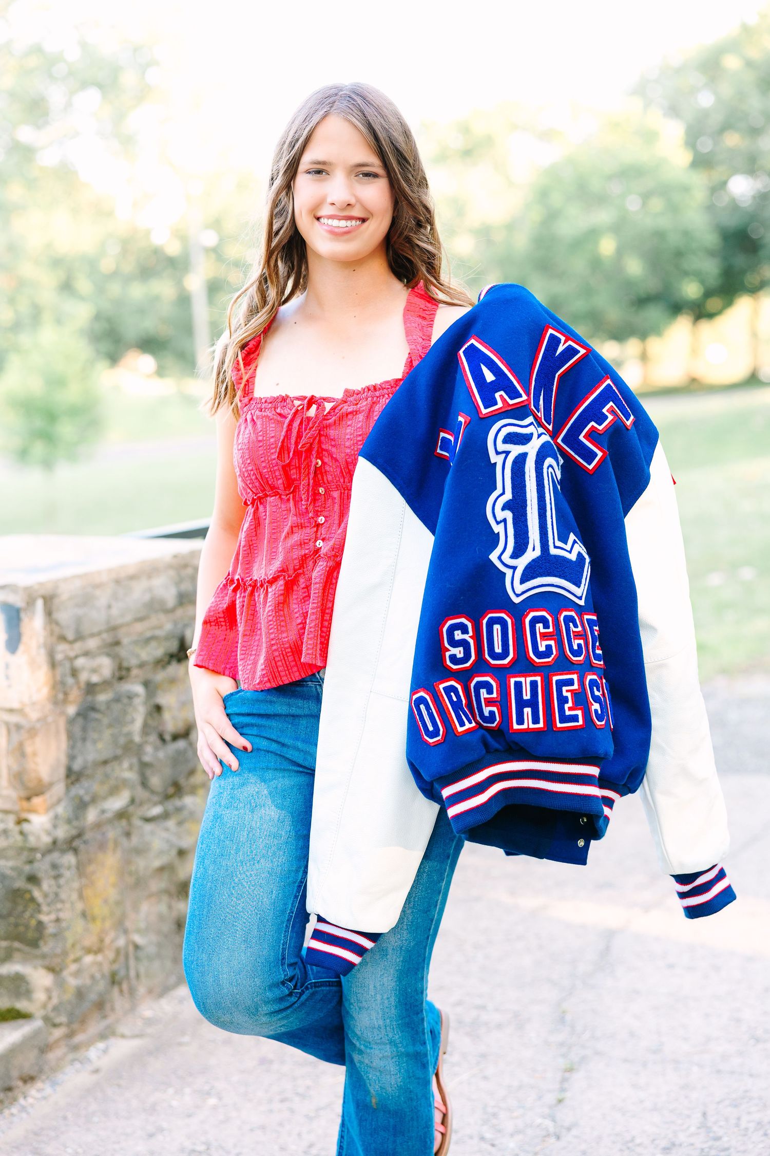 A person wearing a red off-shoulder top and jeans holds a blue and white varsity-style letterman jacket outdoors.