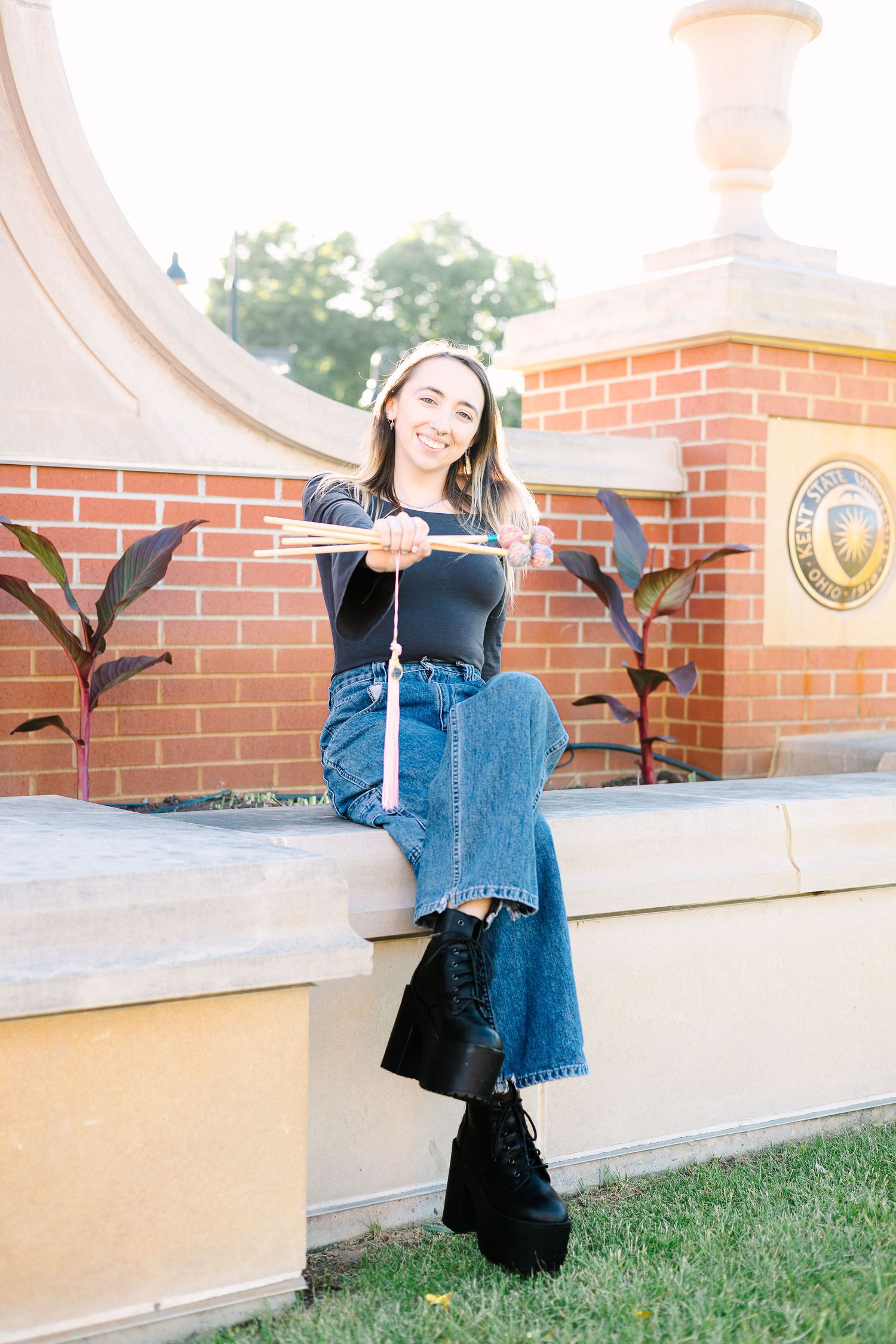 A person in jeans and a black top sits casually on a curved brick wall with decorative plants by a university seal.