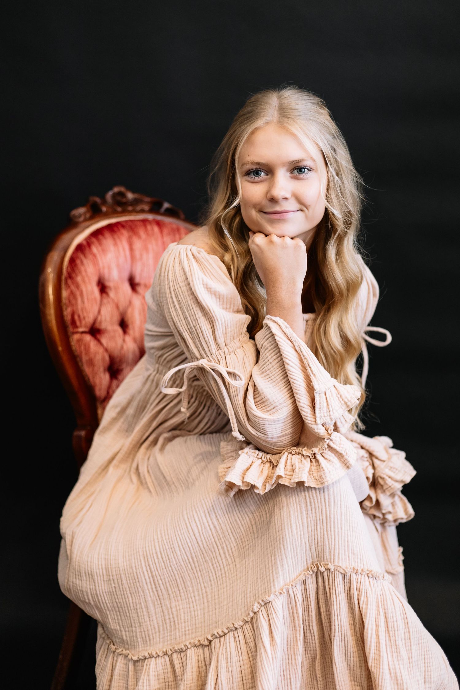 Romantic portrait in cream-colored ruffled dress seated on vintage pink velvet chair.