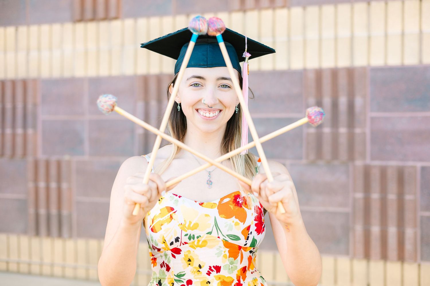 A graduate in a colorful floral dress holds up their drumsticks while wearing a graduation cap.