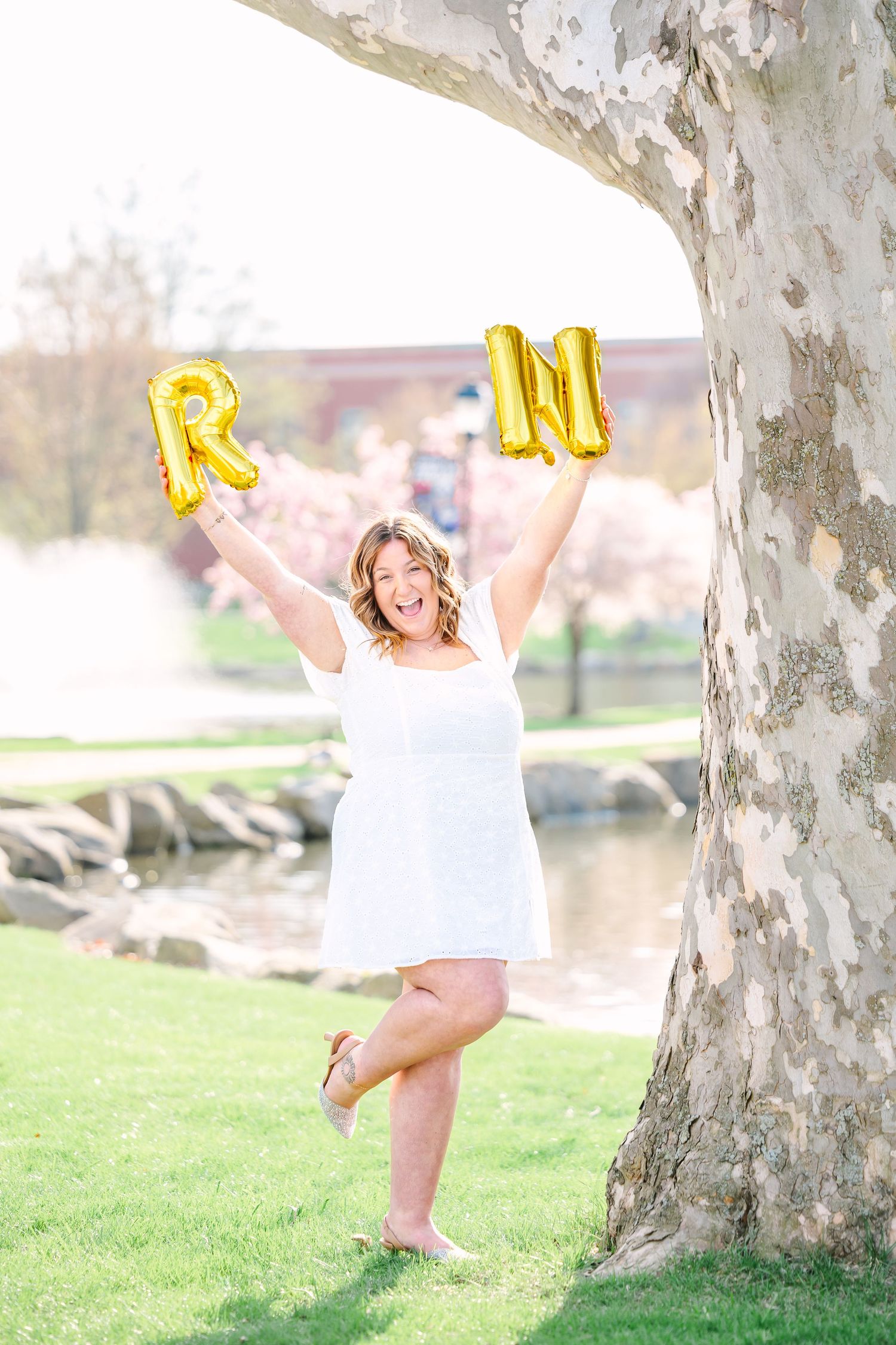 Joyful person in white dress holds gold letter balloons while posing by large tree trunk in sunny outdoor setting.