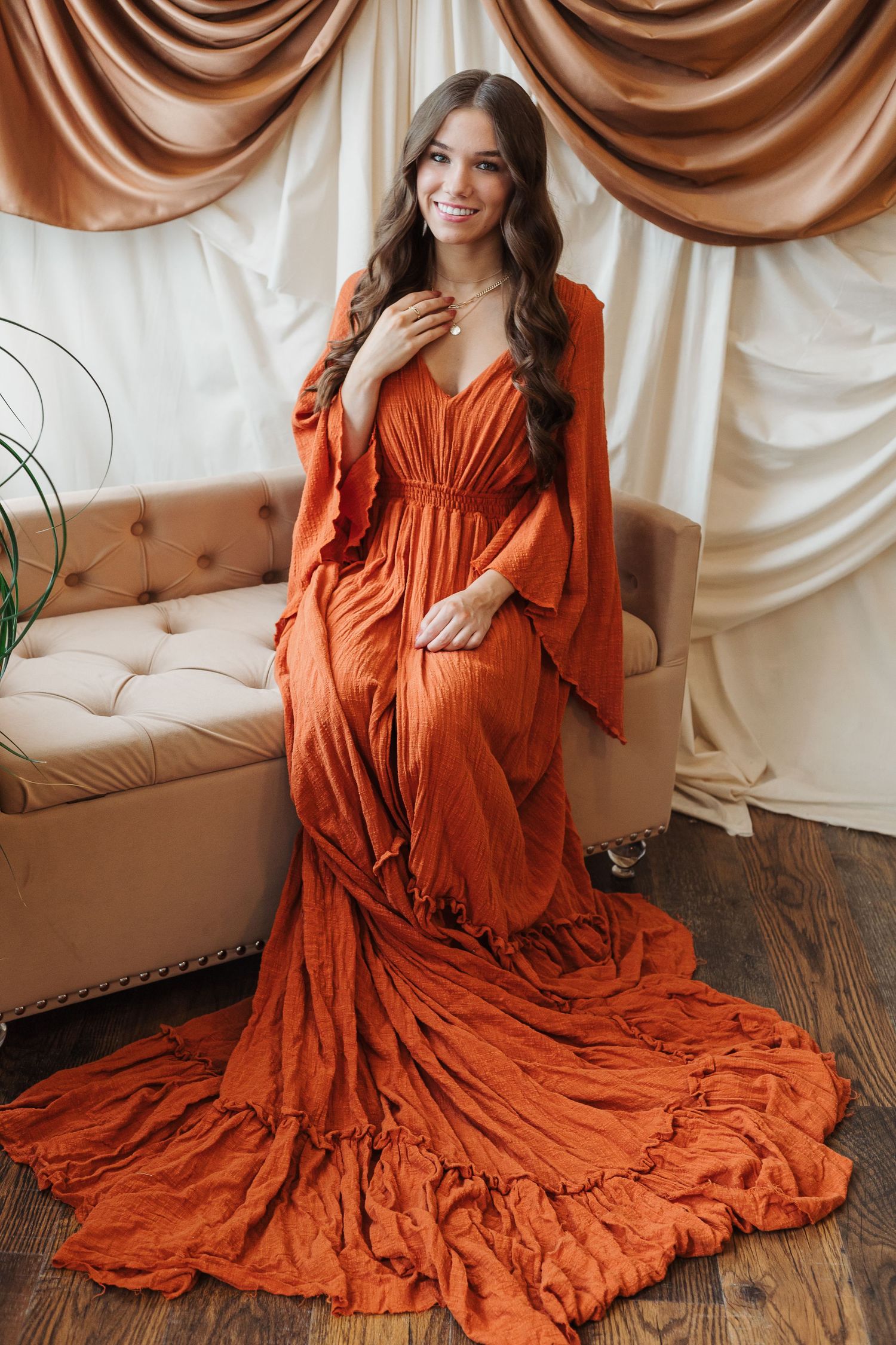 A flowing burnt orange maxi dress with dramatic ruffles and bell sleeves photographed against draped fabric backdrop.