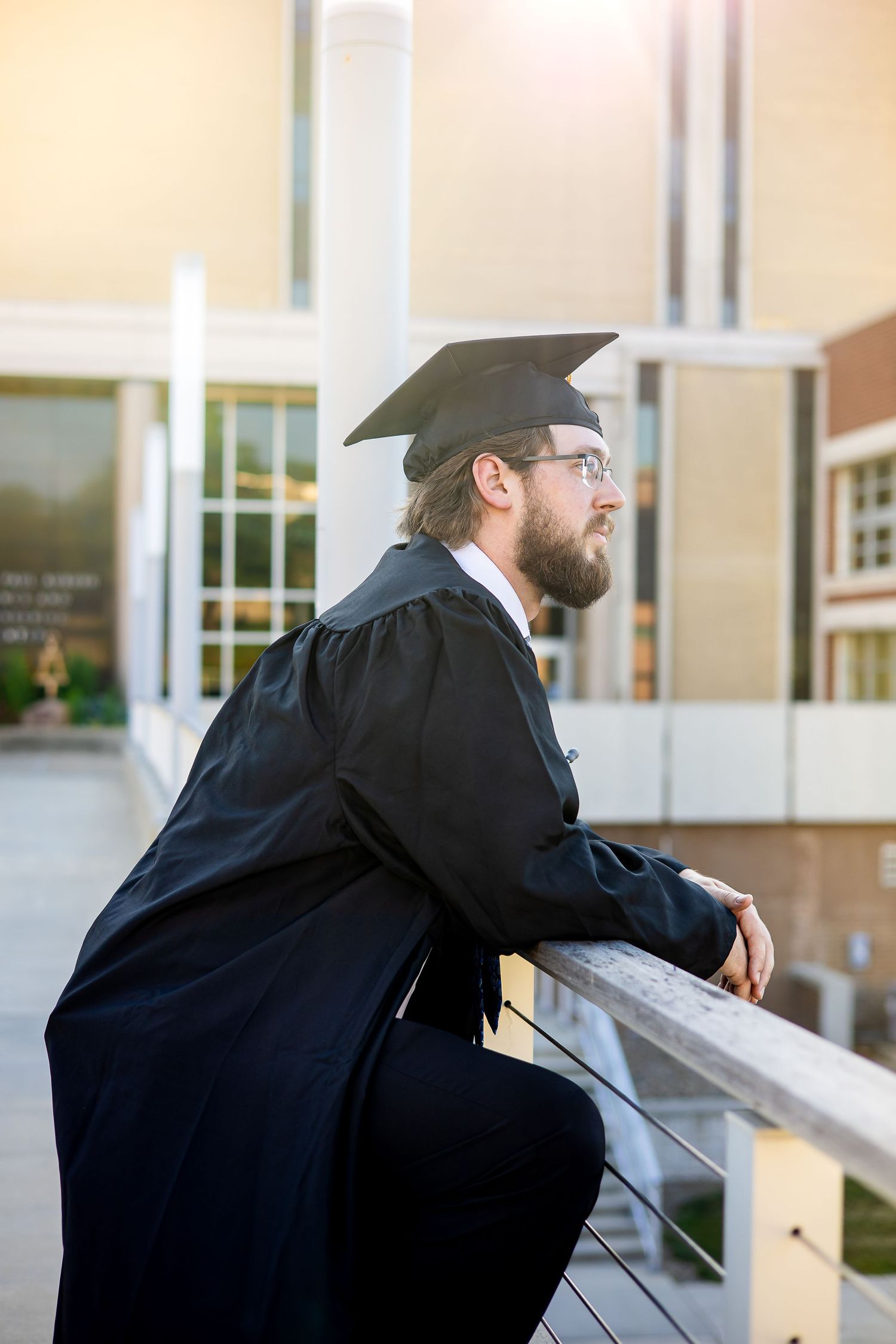 Graduate in academic regalia leans reflectively on outdoor railing at sunset.