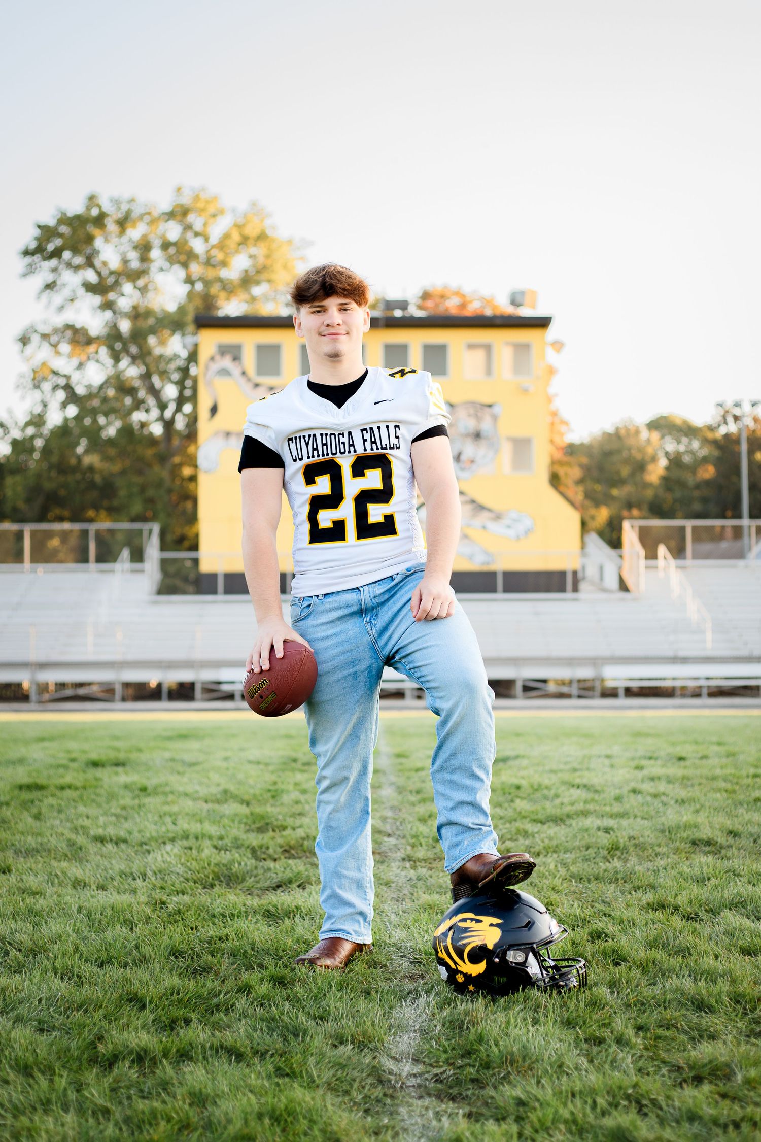 High school football player poses on field in white jersey number 22, light jeans, with football and helmet on grass.