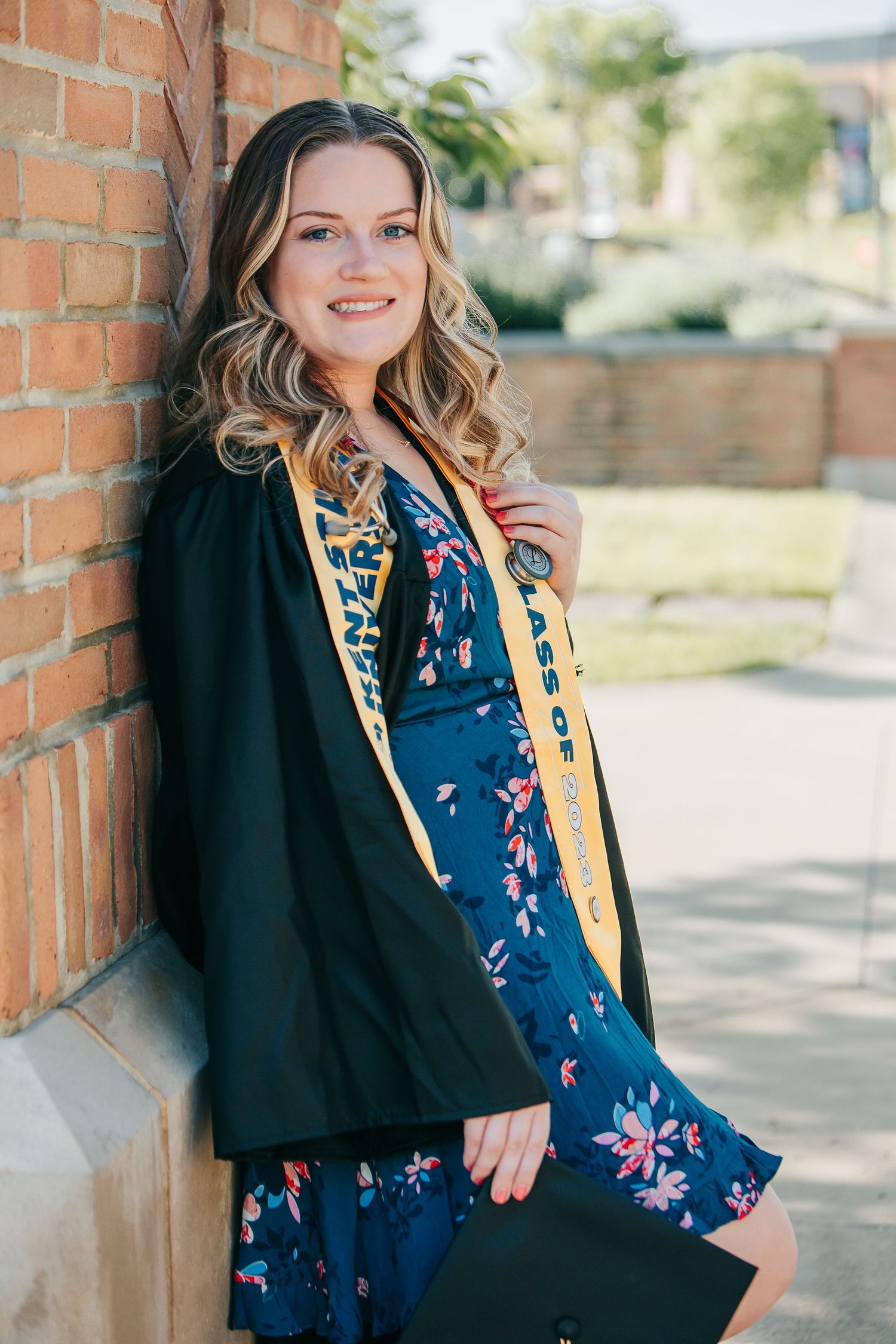 Graduate in a floral blue dress and black blazer poses against brick wall during outdoor photoshoot.